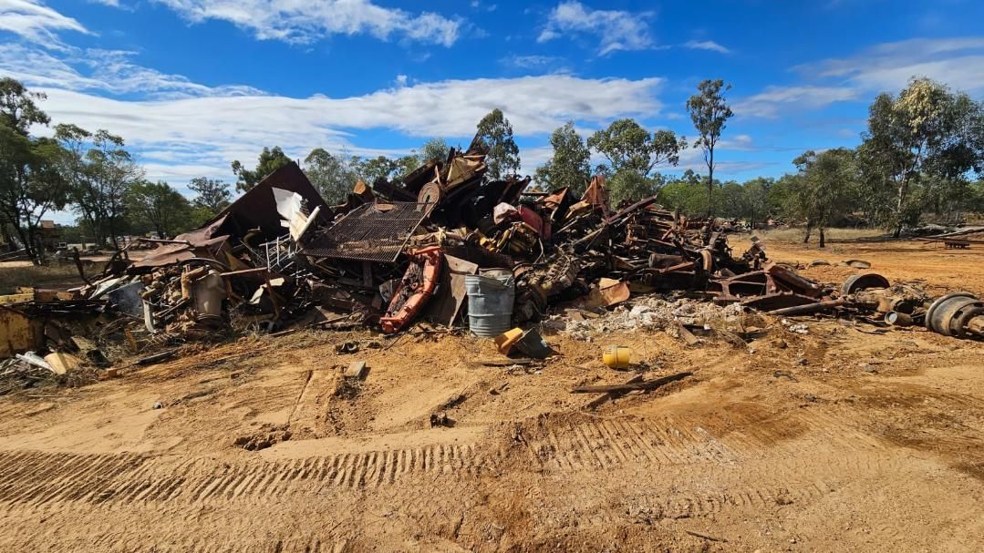 A Pile of Charred Metal and Debris on a Sandy Field Under a Blue Sky With Trees — NPX Solutions In Gympie, QLD