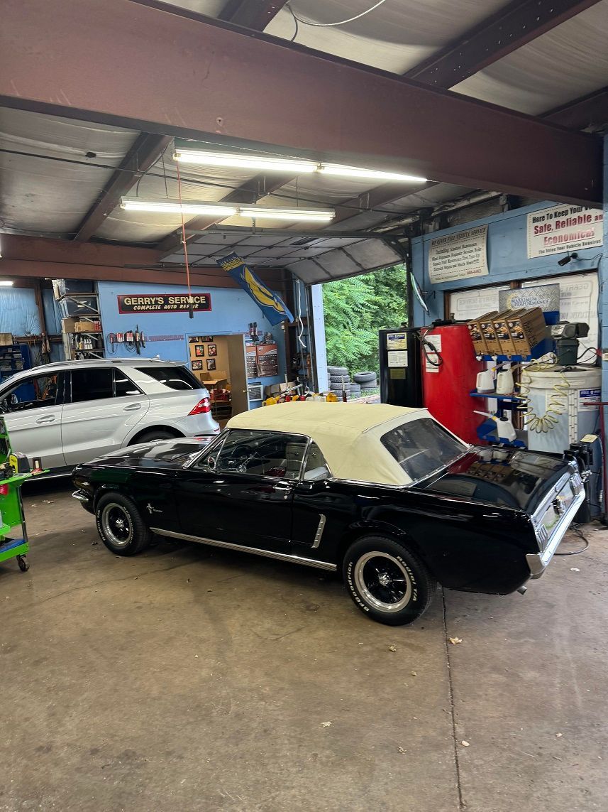 Black convertible Mustang with white top in a repair shop, white SUV in the background.