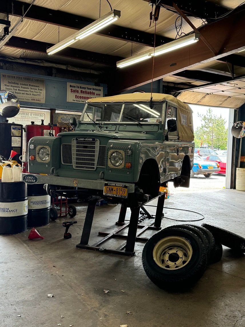 A Land Rover Defender on a lift in a garage. It's painted green with a tan top. Tires on the floor.