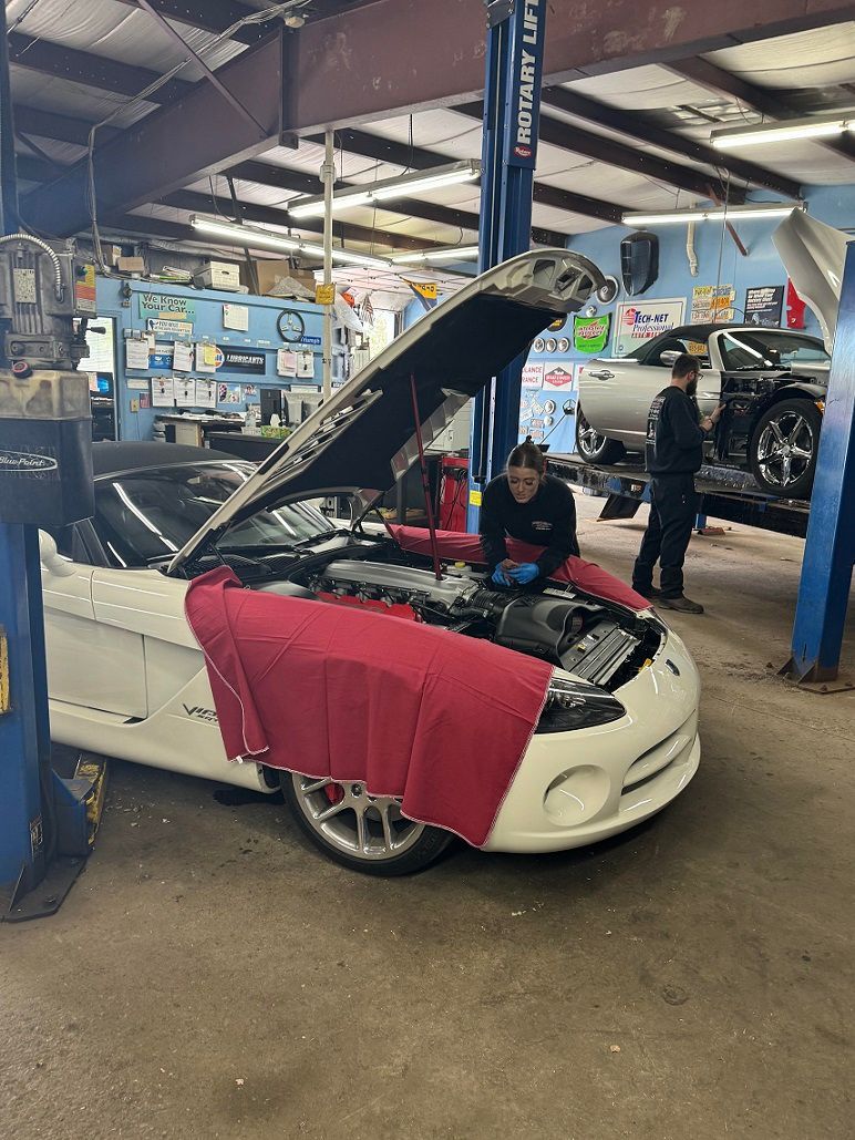 White sports car with hood up in a repair shop, two mechanics working on the engine.