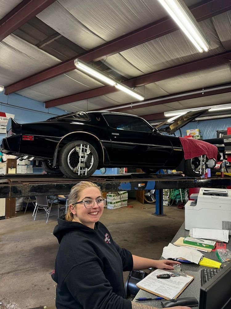 Woman at desk smiles in auto repair shop with black classic car on lift.