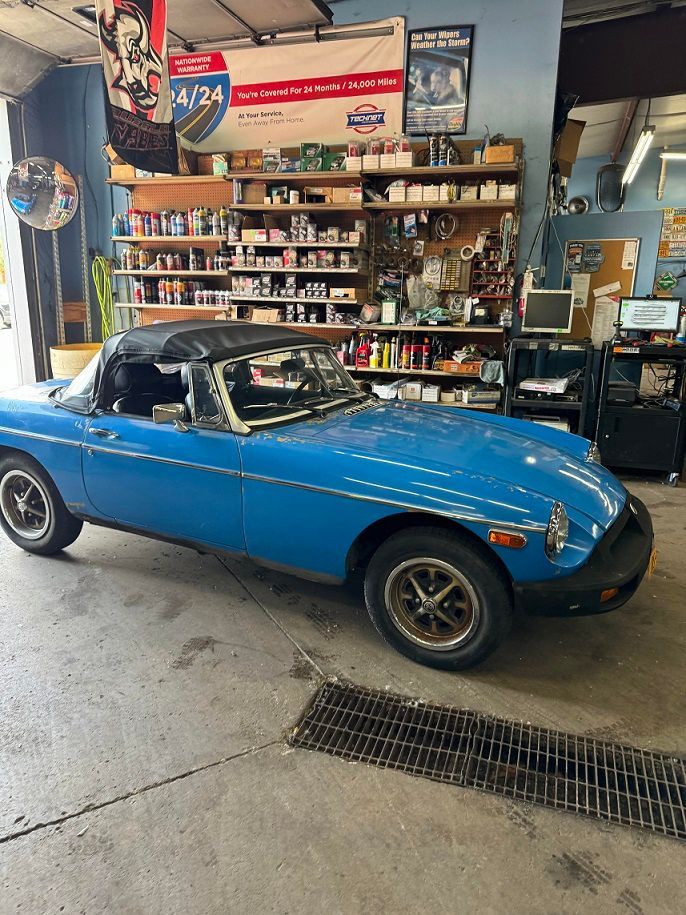 Blue convertible car inside a mechanic's shop, black top down. Shelves with parts visible in background.