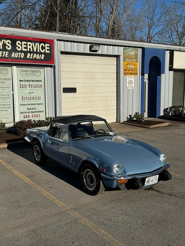 Light blue Triumph Spitfire parked in front of an auto repair shop with a white garage door.