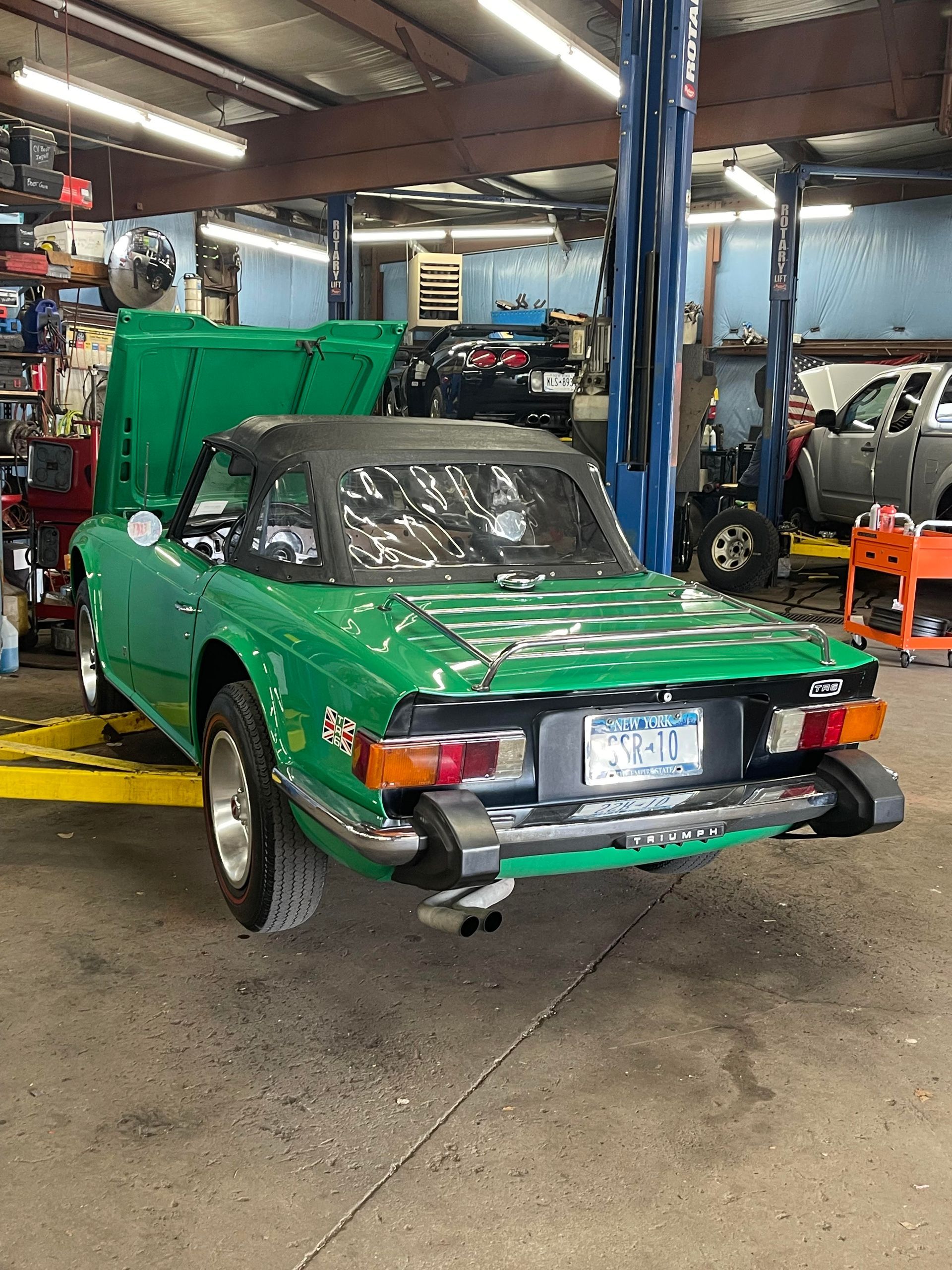 Green Triumph TR7 convertible in a garage, hood open, on a car lift.