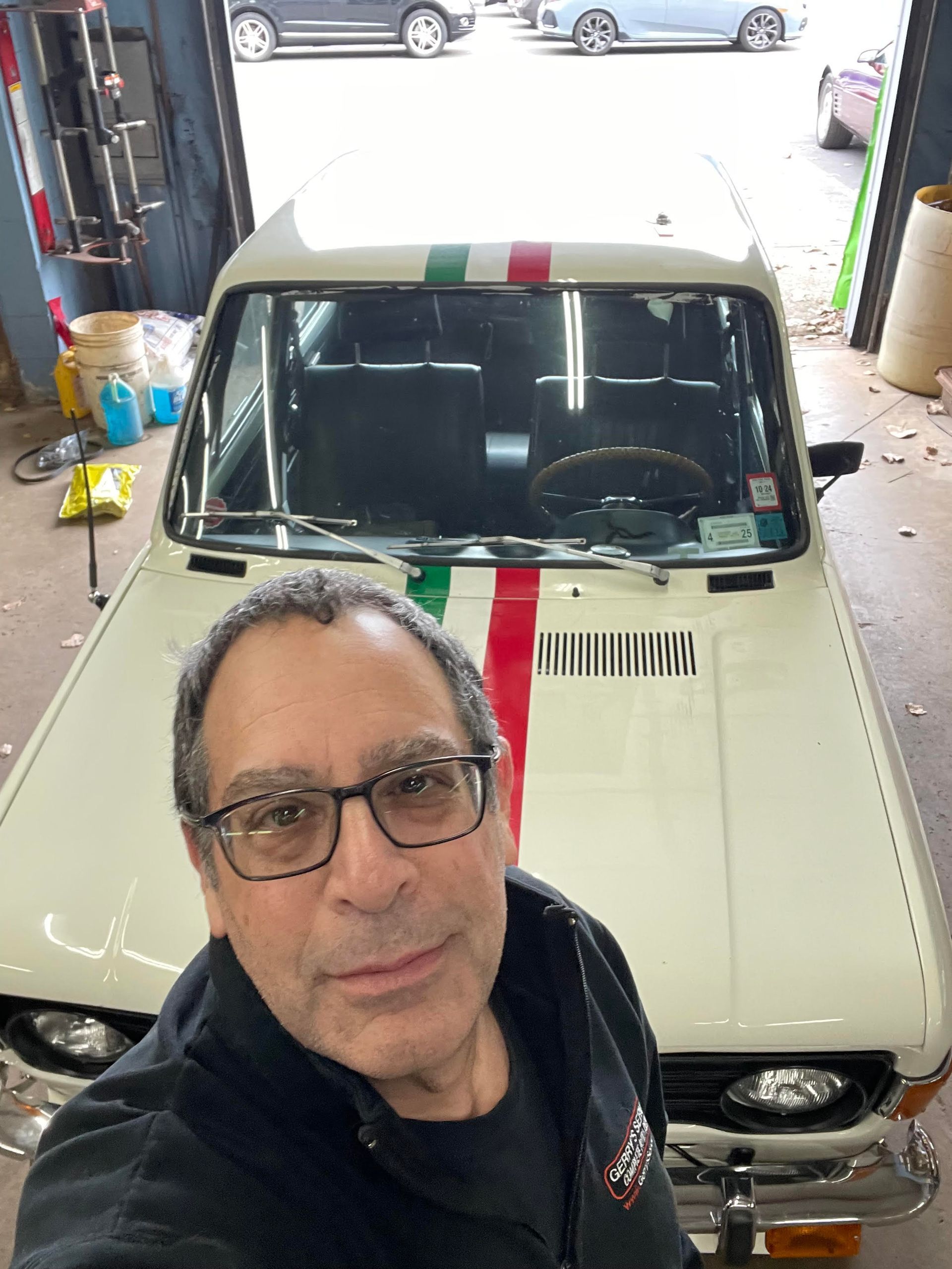 Man in glasses with classic white car with Italian flag stripe on hood, in auto shop.