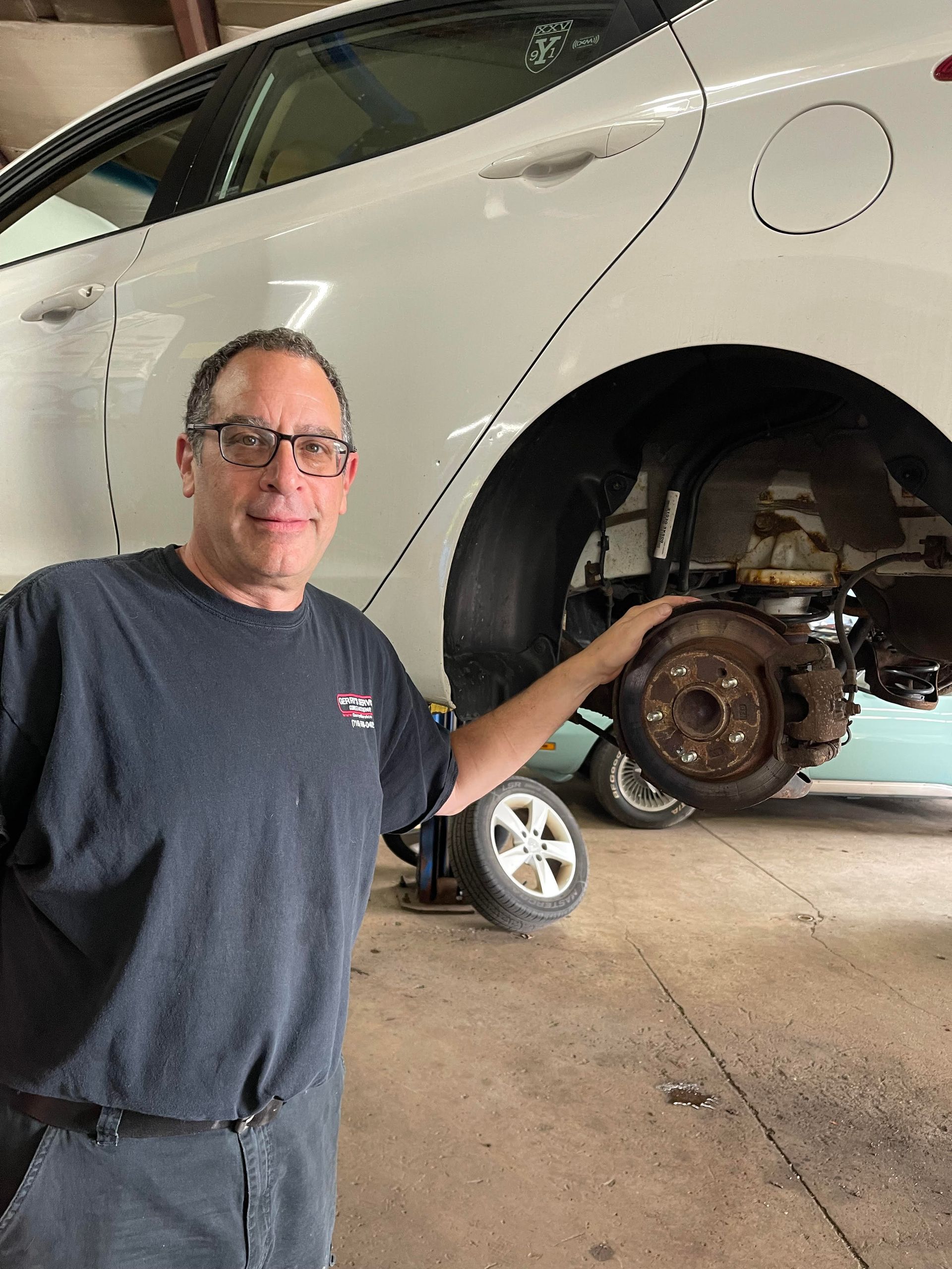 Mechanic holding car brake rotor, standing next to a white car in a garage.