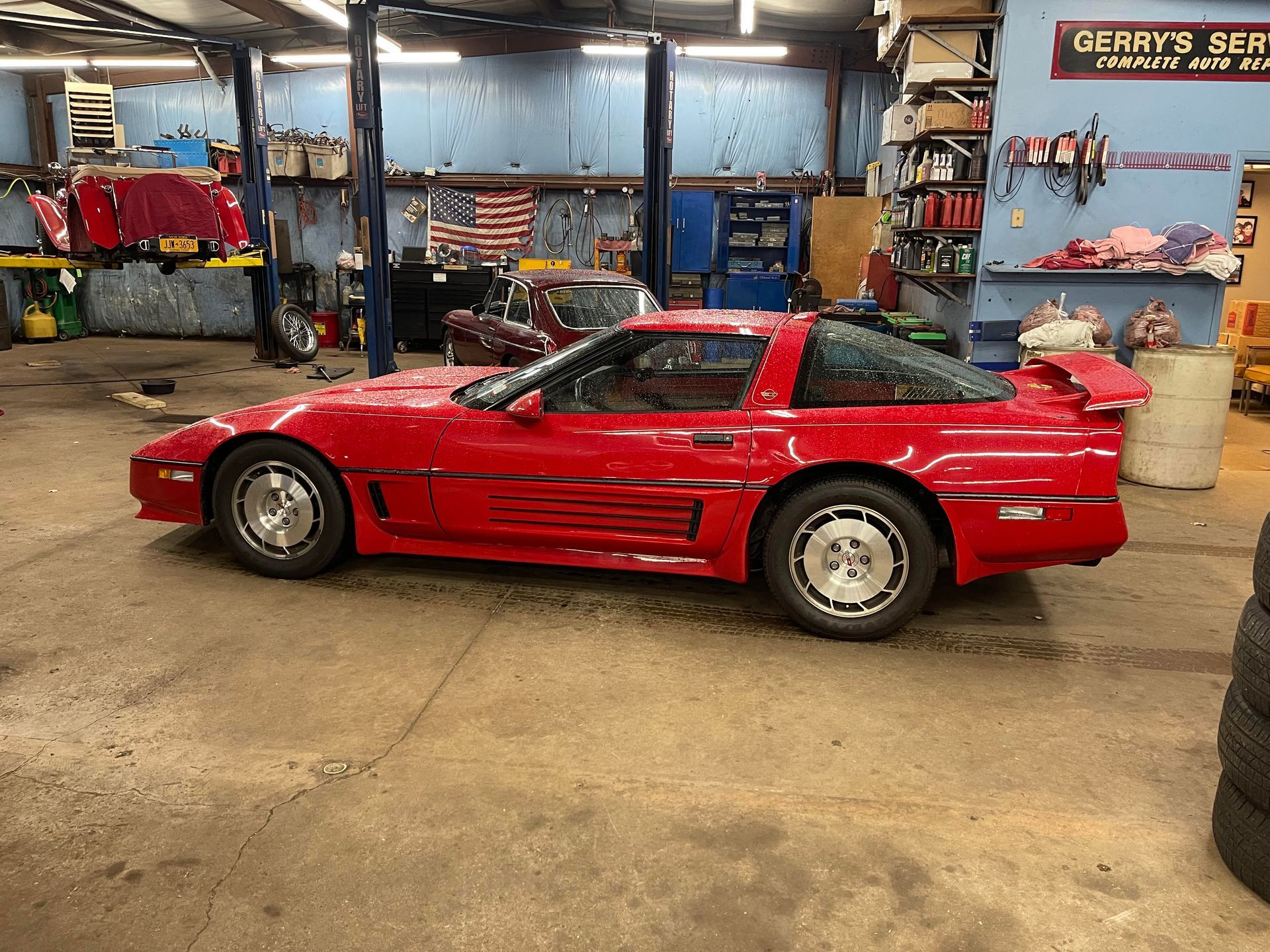 Red Chevrolet Corvette in a cluttered garage, possibly being worked on.