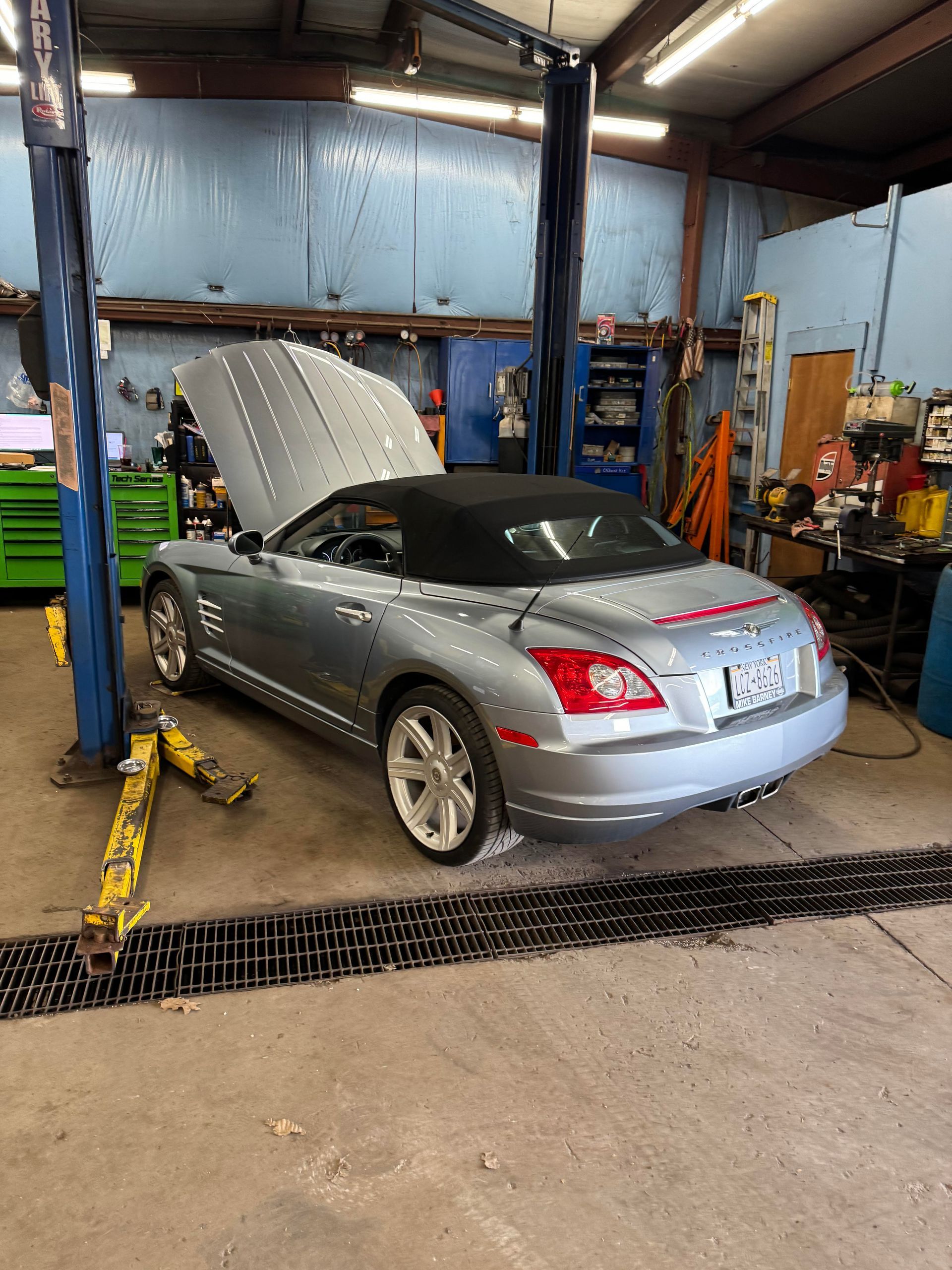 Silver convertible car in a repair shop with hood open, on a lift. Black roof, blue walls, tools in background.