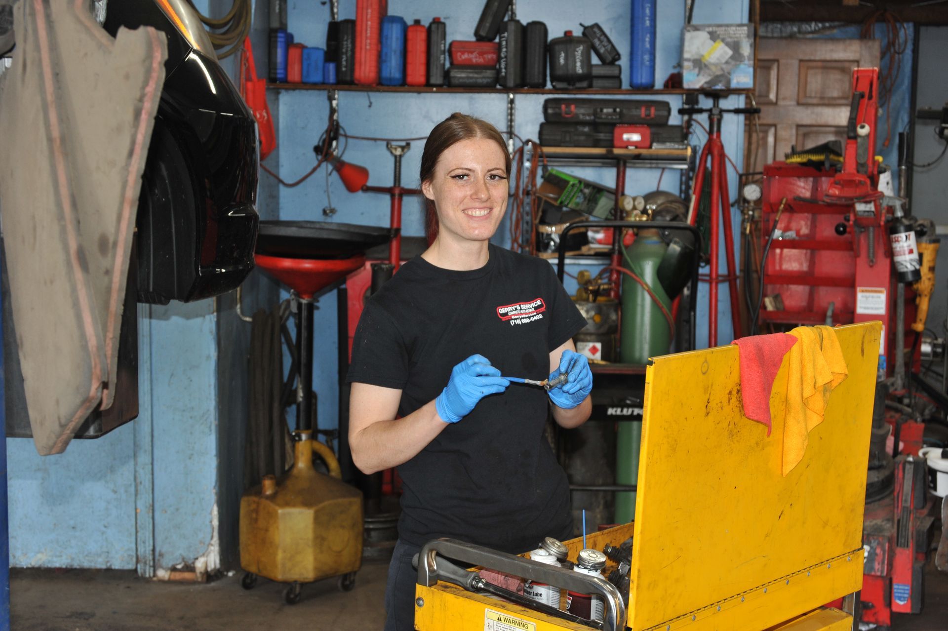 Woman in auto shop holding a wrench, smiling. Yellow tool cart, blue gloves, setting with tools.