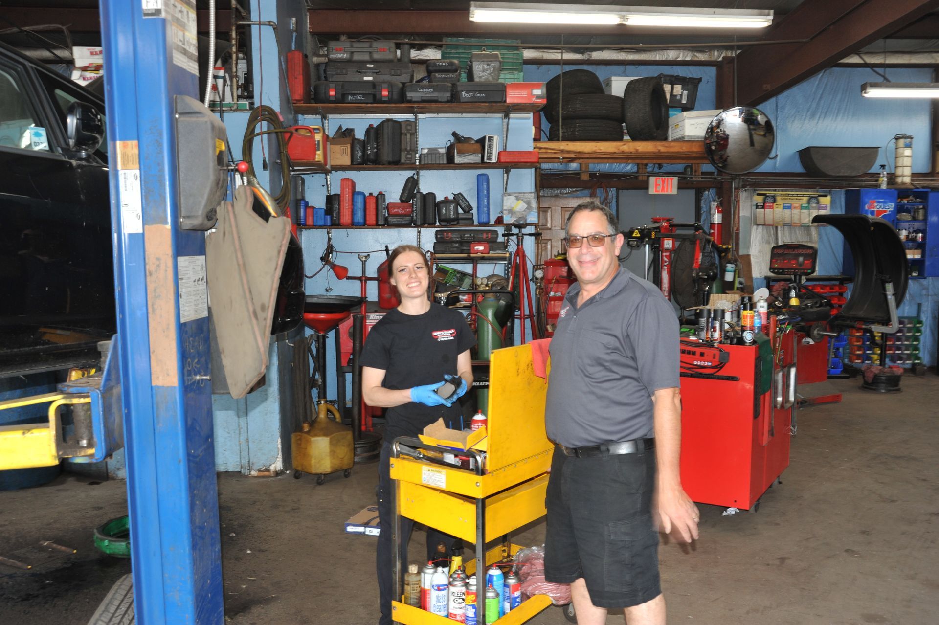 Two people in a garage: a woman in blue gloves and a man smiling; tools and equipment visible.