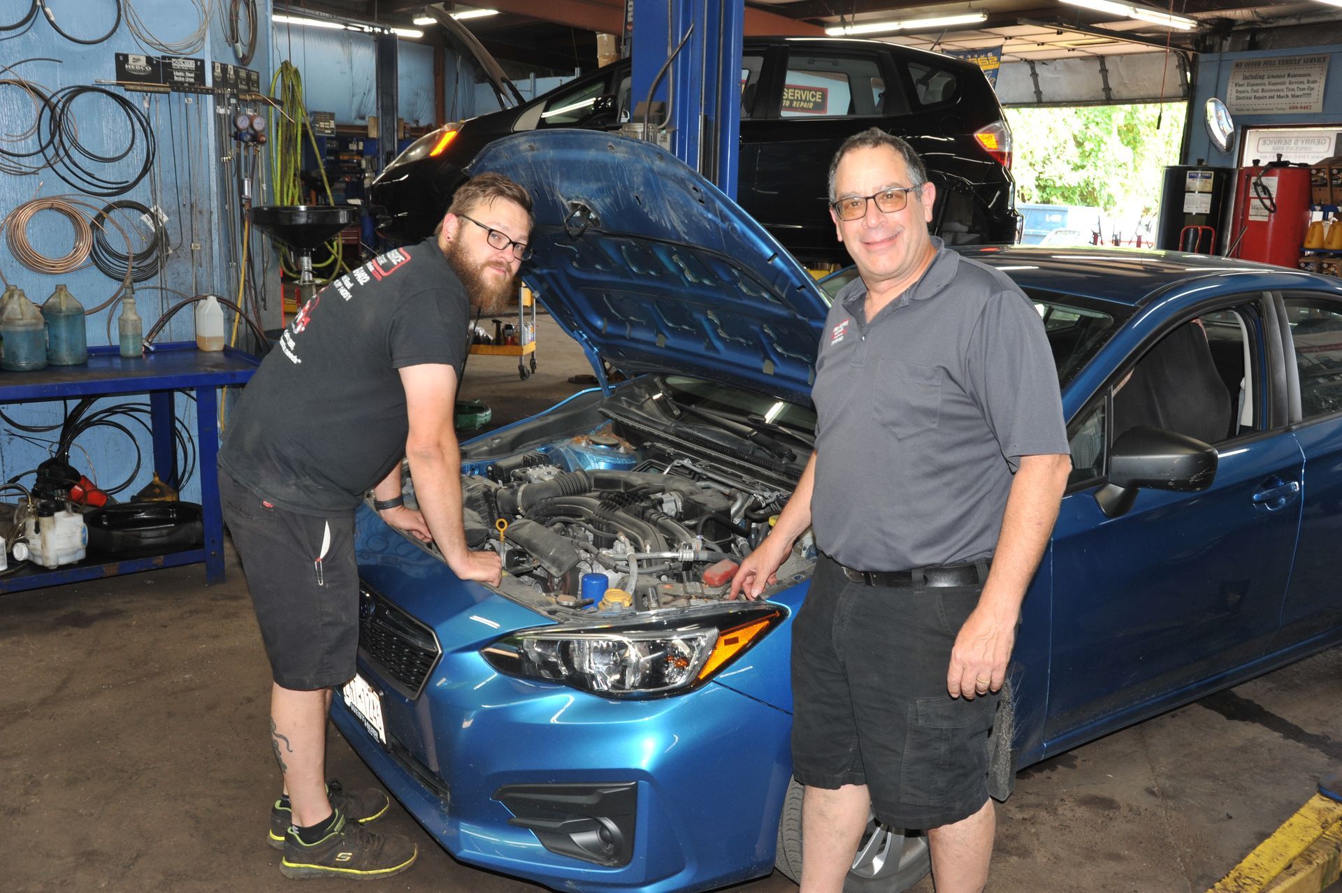 Two men examine a blue car's engine in a garage. One points, the other looks on.