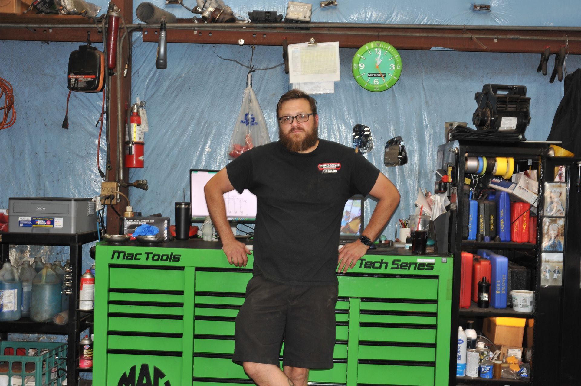 Man in a black t-shirt and shorts, leaning on a green Mac Tools toolbox in a cluttered garage.