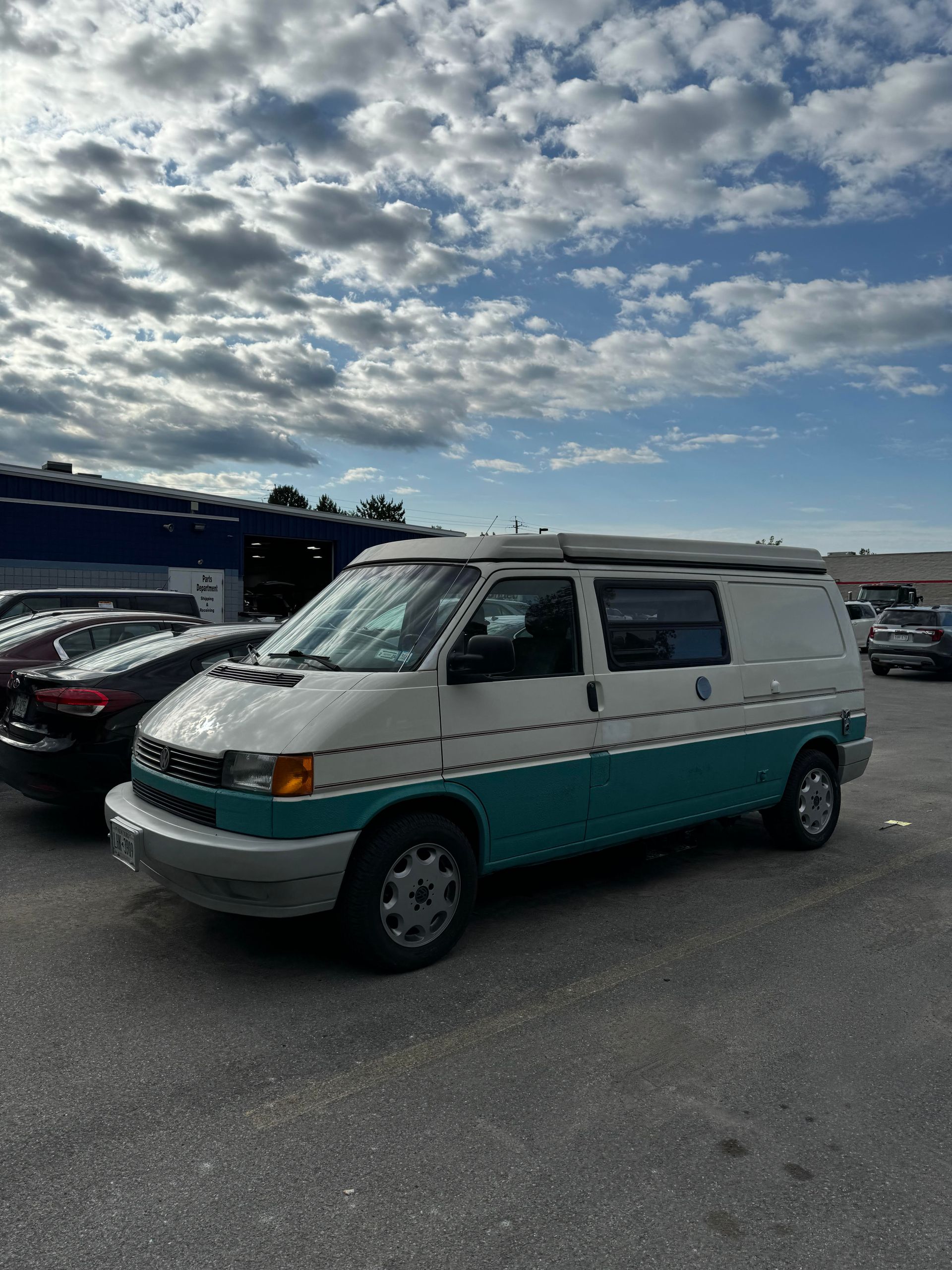 White and teal Volkswagen Eurovan camper van parked in a lot, cloudy sky overhead.