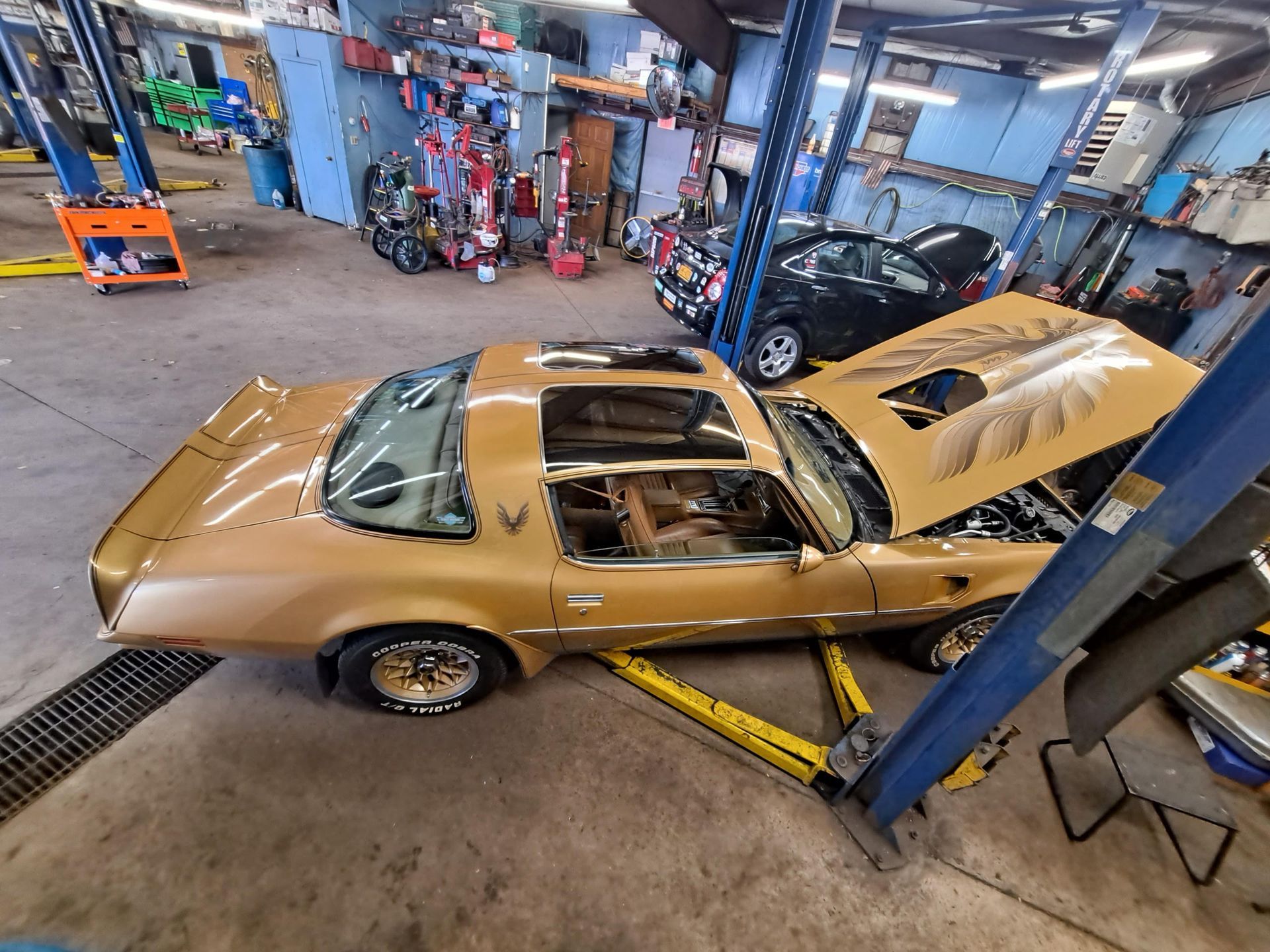 Gold Pontiac Firebird on a lift in a repair shop. Hood open.