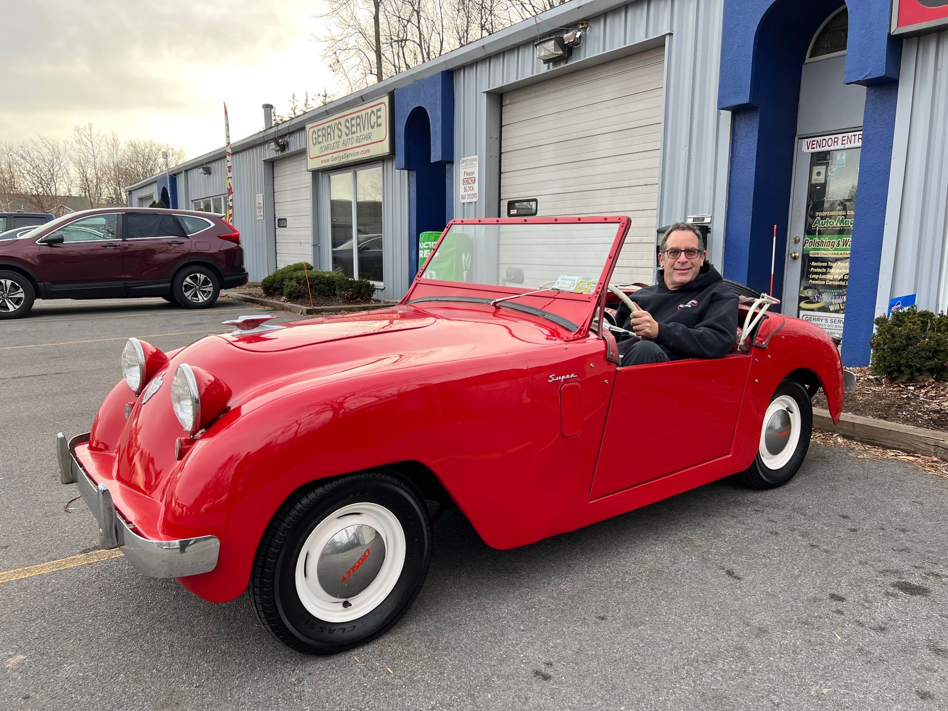 Man in a red vintage car, parked in front of a blue garage.