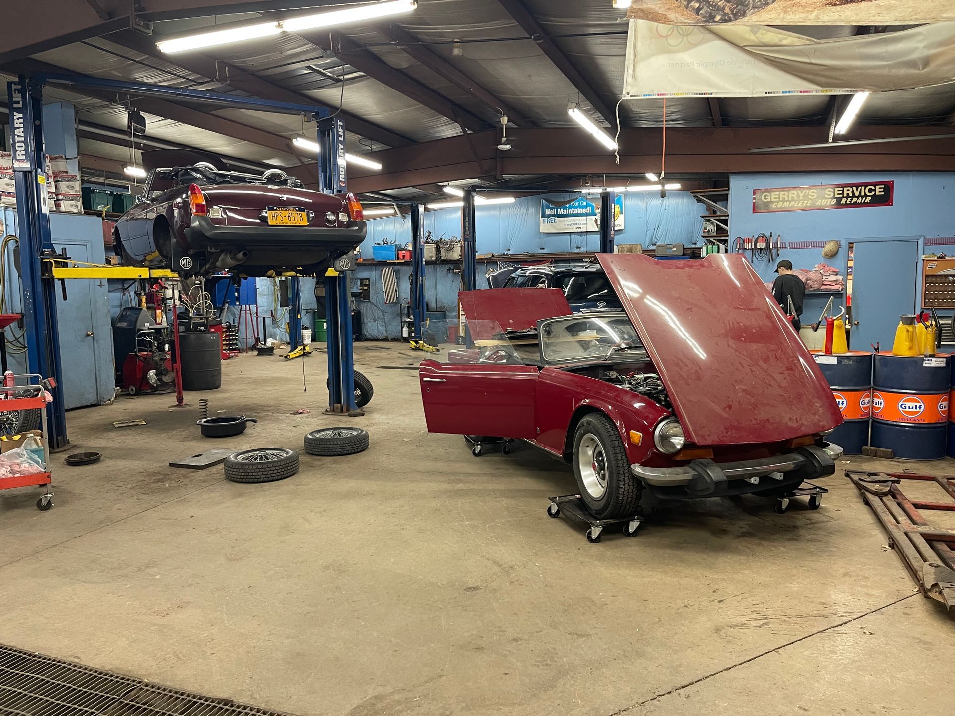 Auto repair shop with two cars in various states of disassembly. One car raised on a lift, another with hood open.