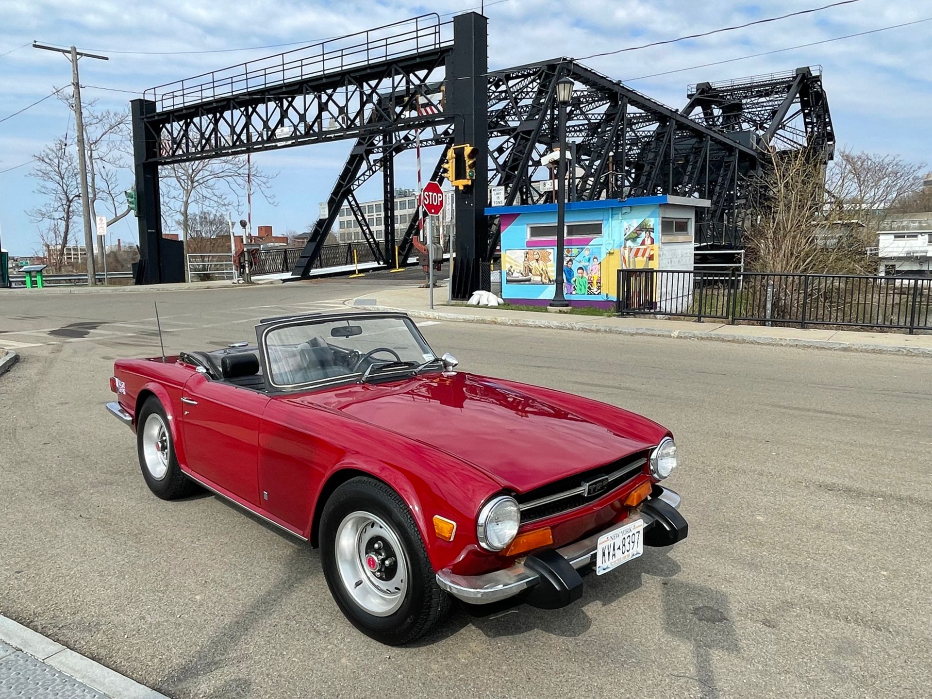 Red convertible sports car parked near a black metal bridge and colorful building on a sunny day.
