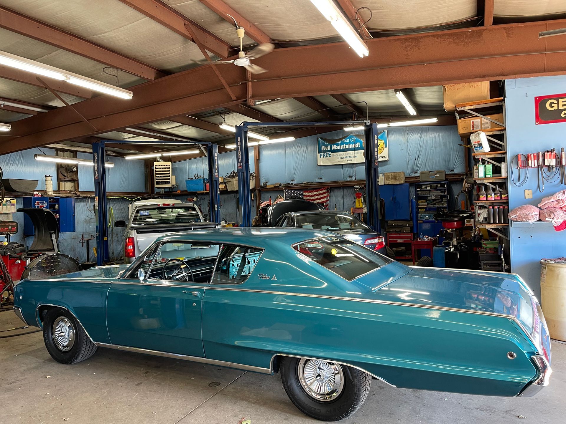 Teal classic car inside a cluttered auto repair shop, with car lifts and tools.