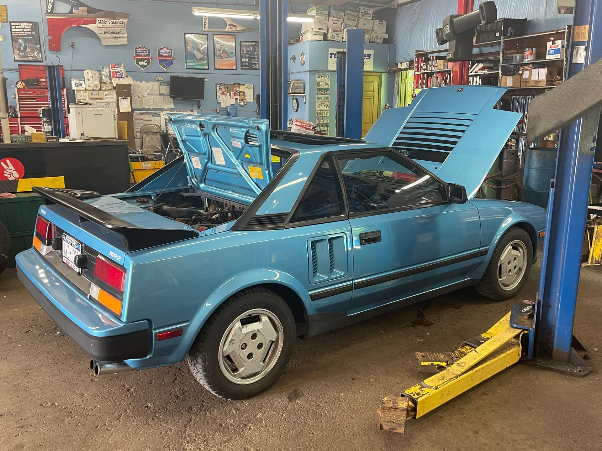 Blue Toyota MR2 in a repair shop with hood and trunk open, on a lift.