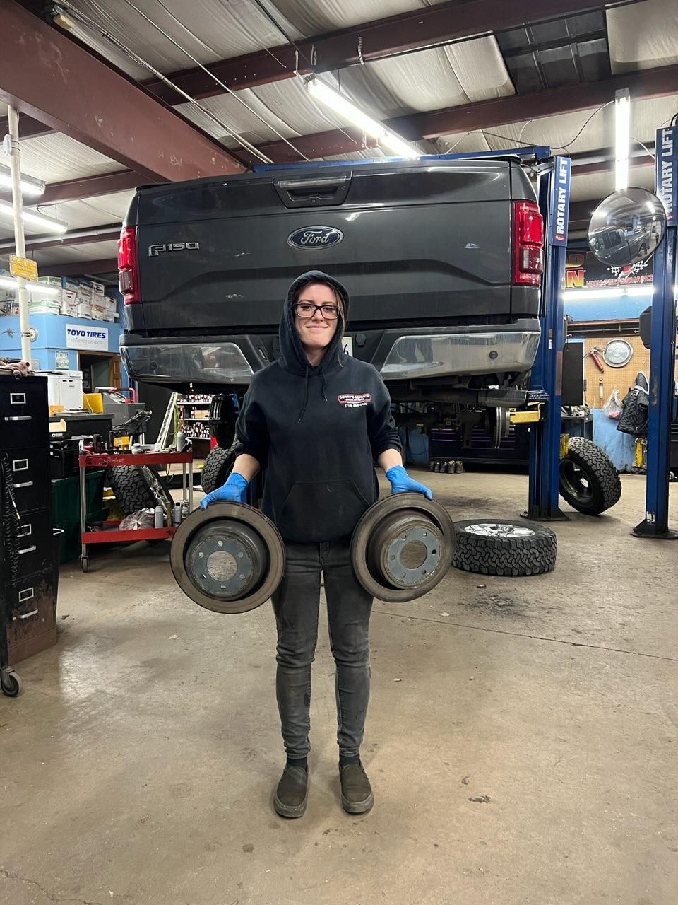 Person holding two brake drums in a garage with a truck elevated on a lift.