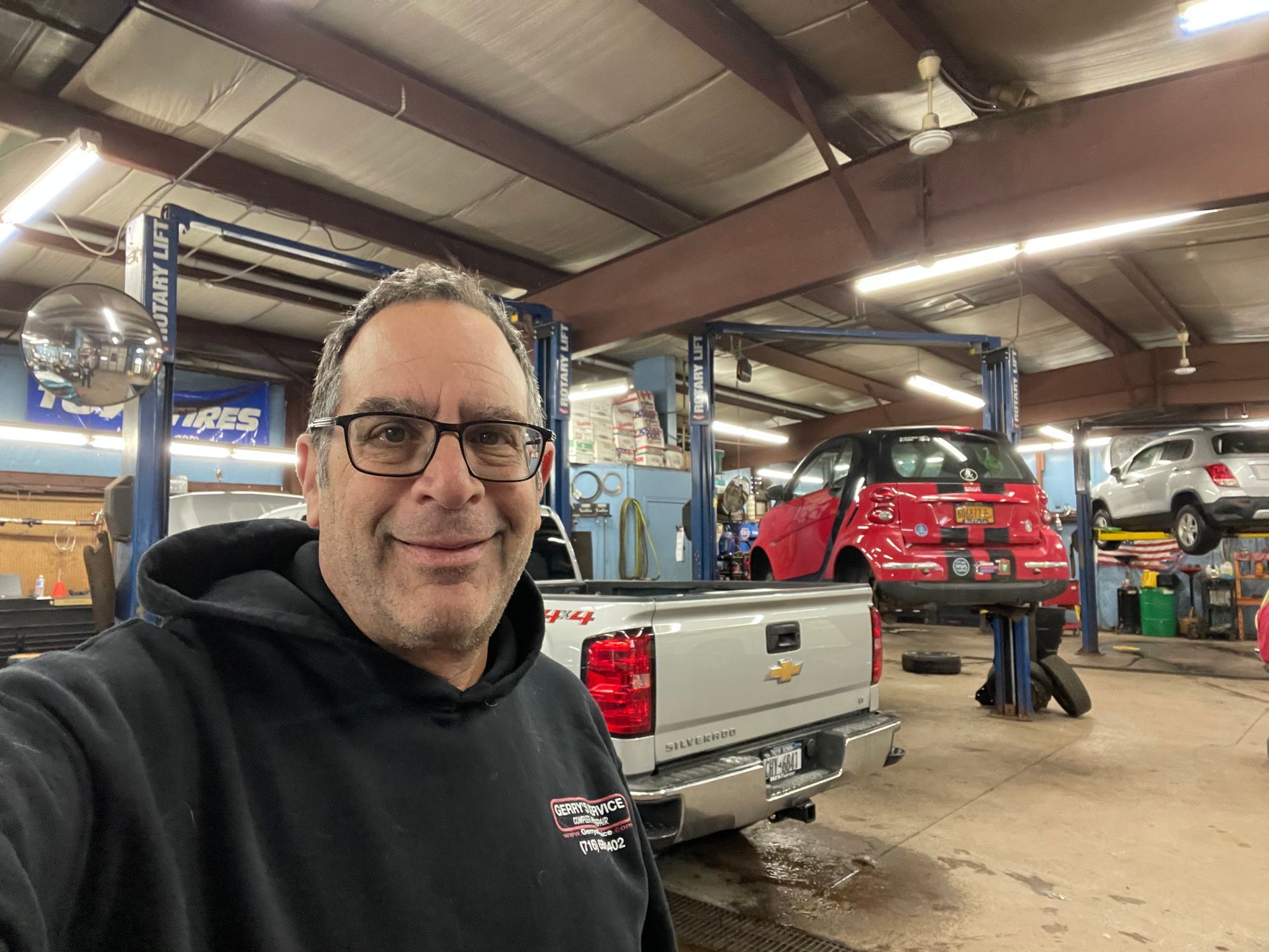Man in a garage smiles at camera. Silver pickup truck in foreground, red car on lift in background.