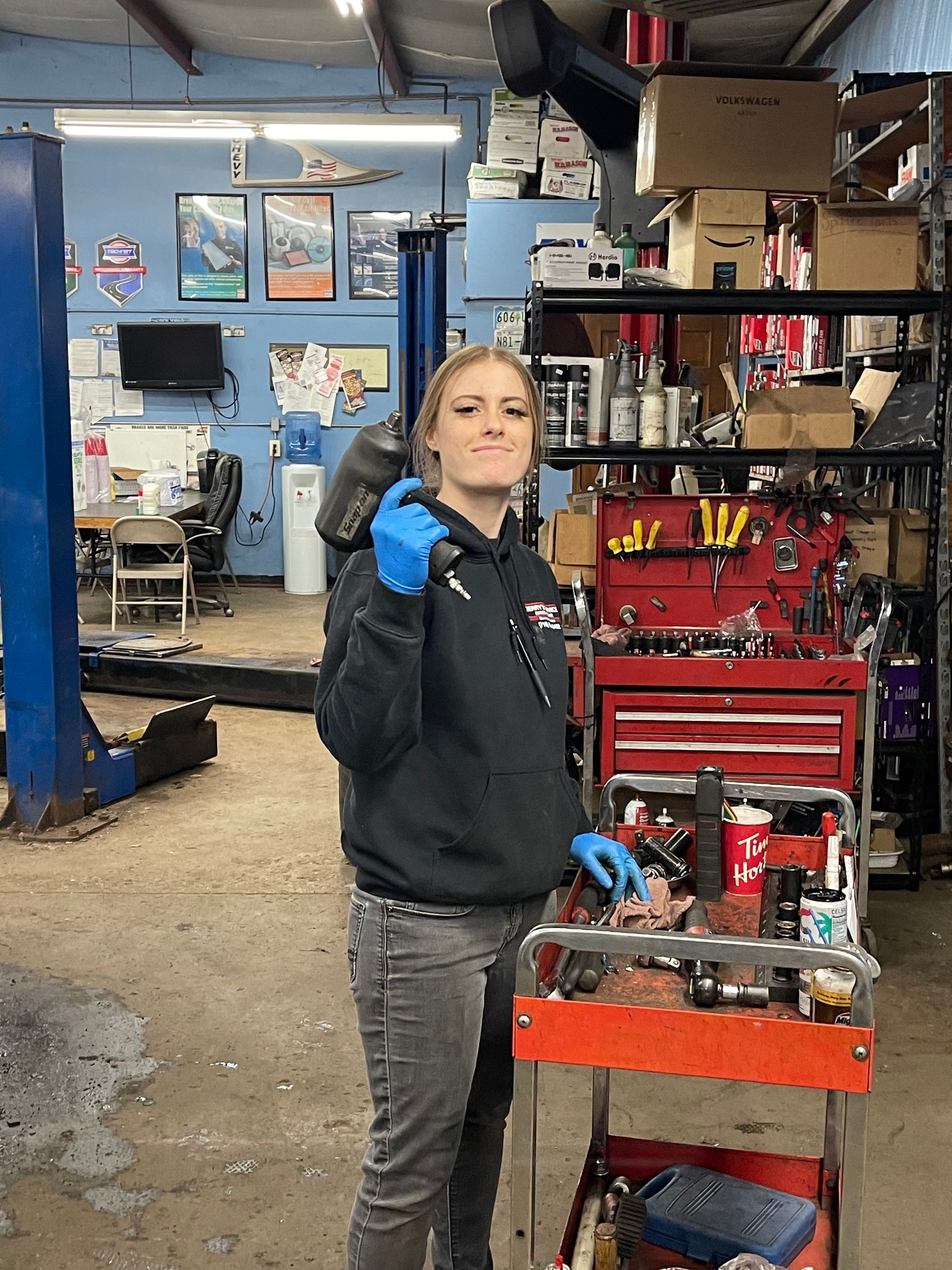 Woman in auto shop holding a tool, smiling, wearing a black hoodie and gloves; tools and parts visible in the background.