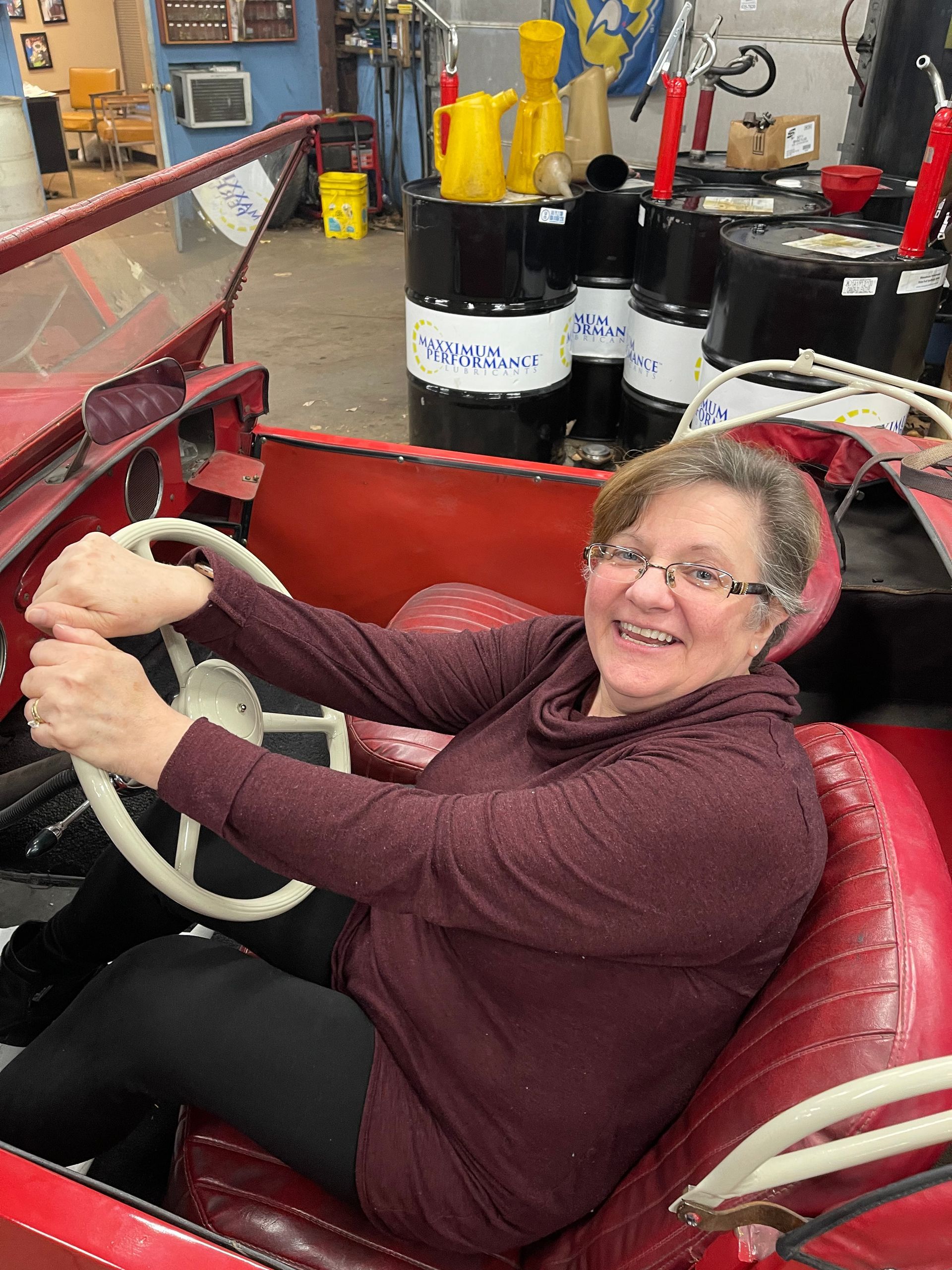 Woman smiling, driving a small red toy car indoors with oil barrels in the background.
