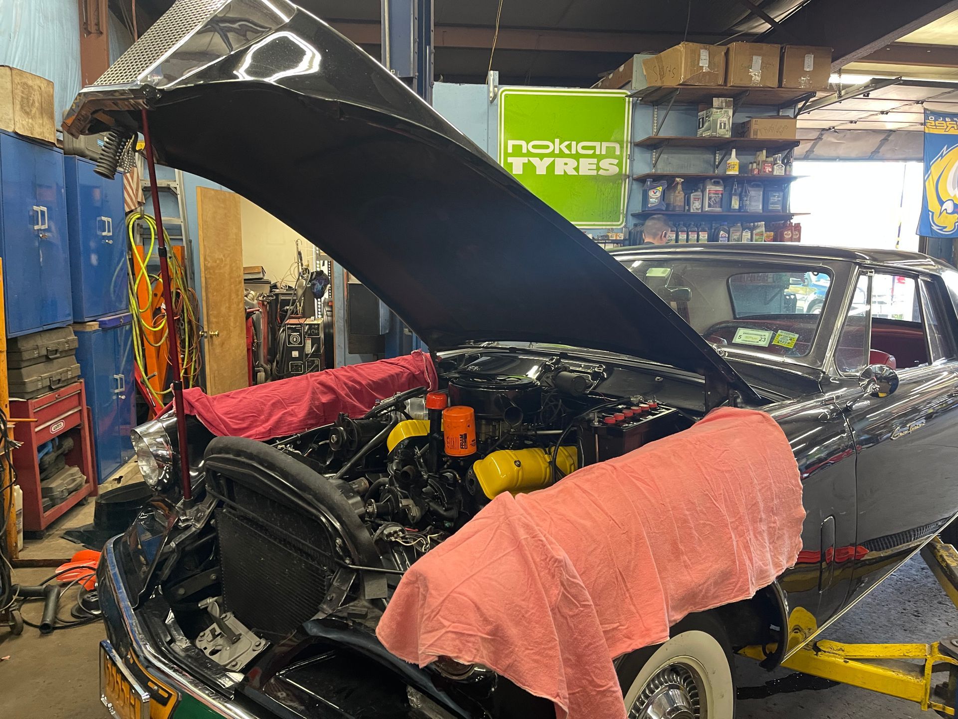 Black car with open hood in a garage. Pink cloths protect fenders. Engine visible with yellow parts.