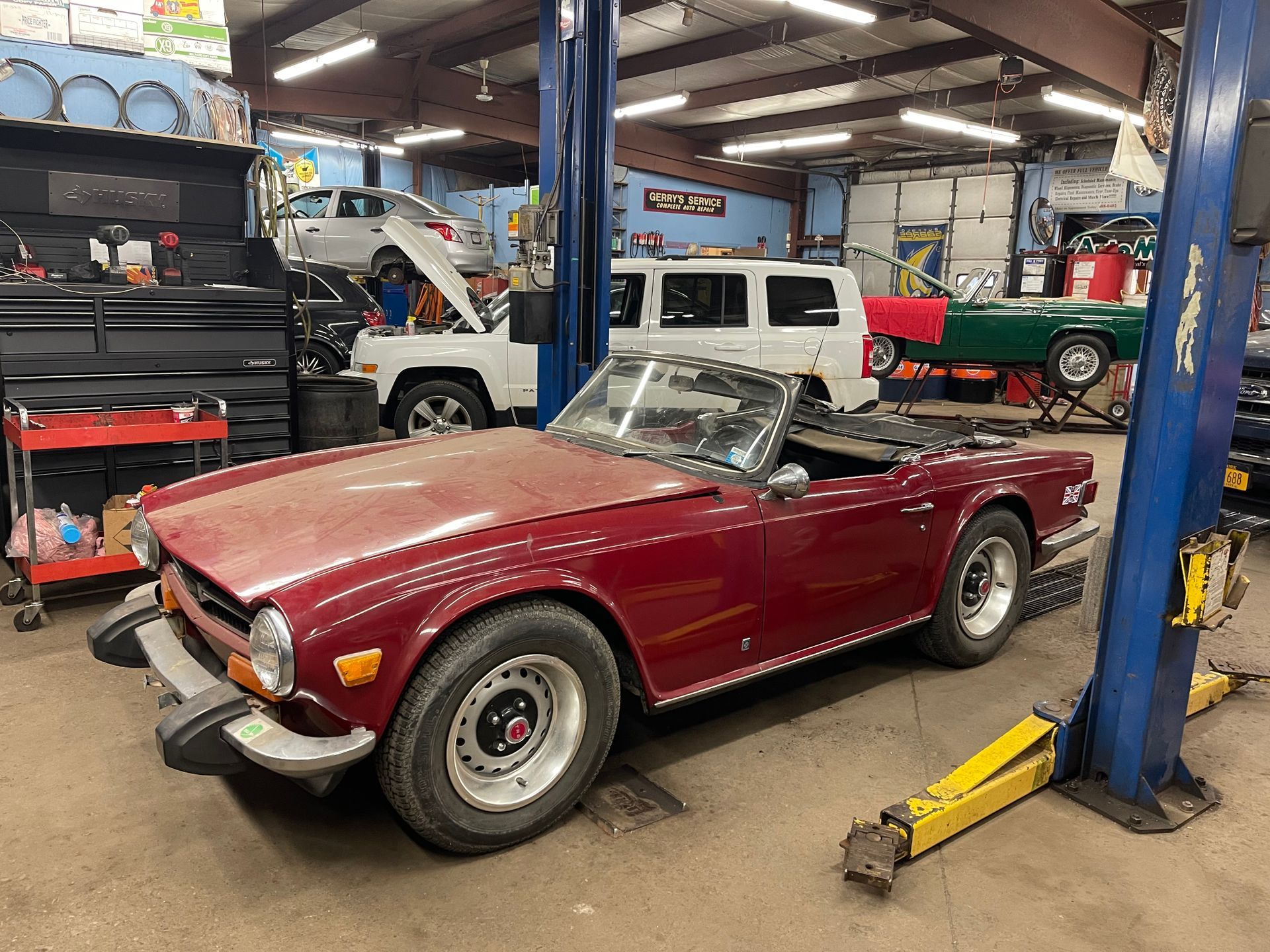 Maroon convertible sports car in a cluttered auto repair shop, hood up, surrounded by other vehicles.