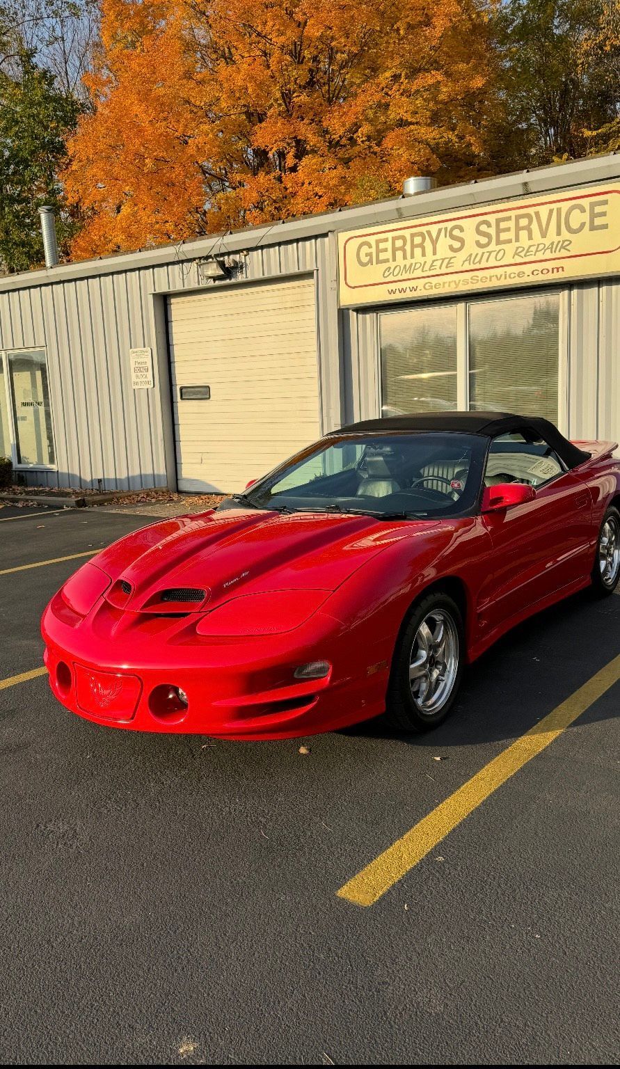 Red convertible Pontiac Firebird parked outside a service garage with fall foliage.