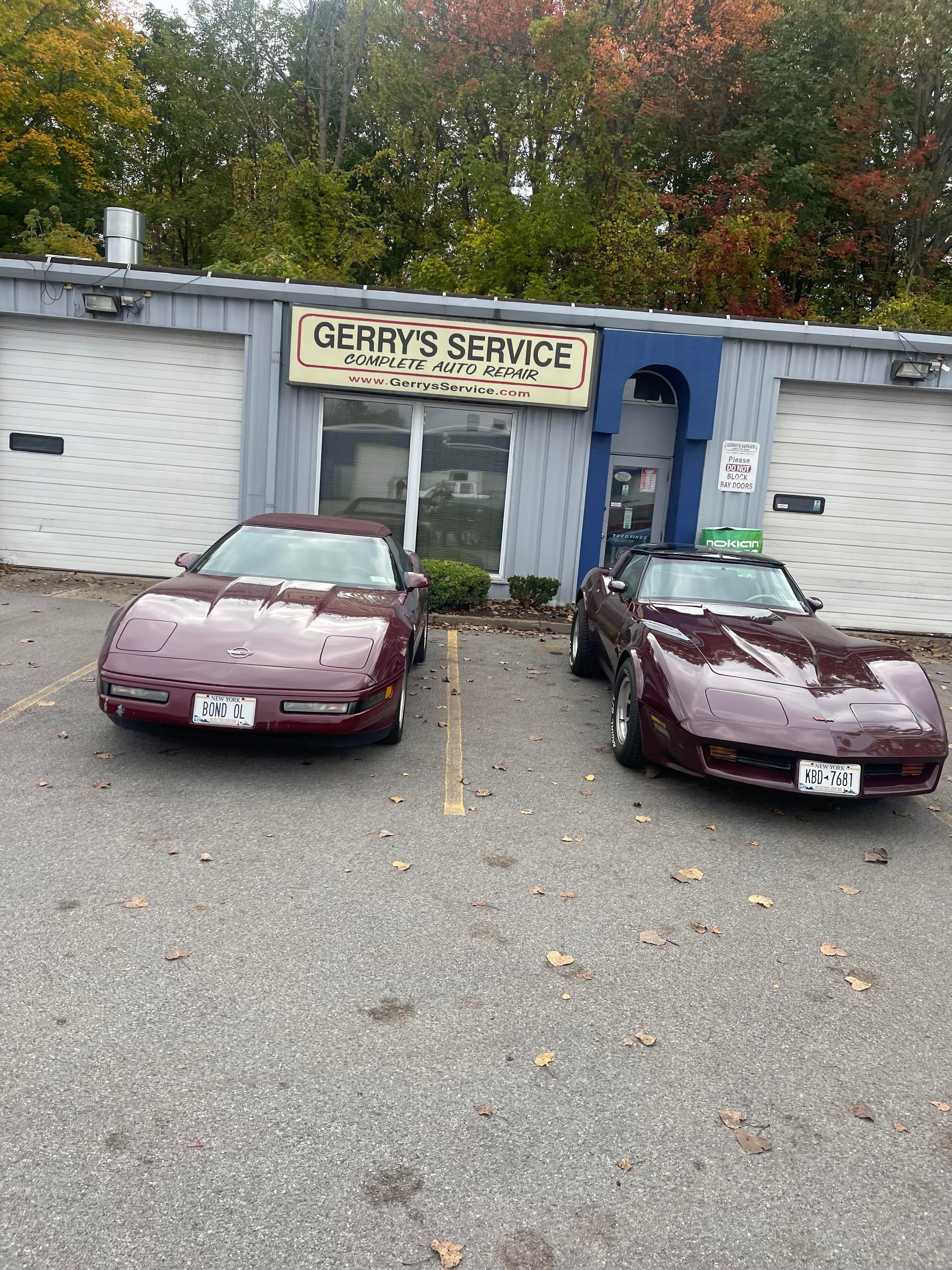 Two maroon Corvettes parked outside Gerry's Service garage with fall foliage in the background.