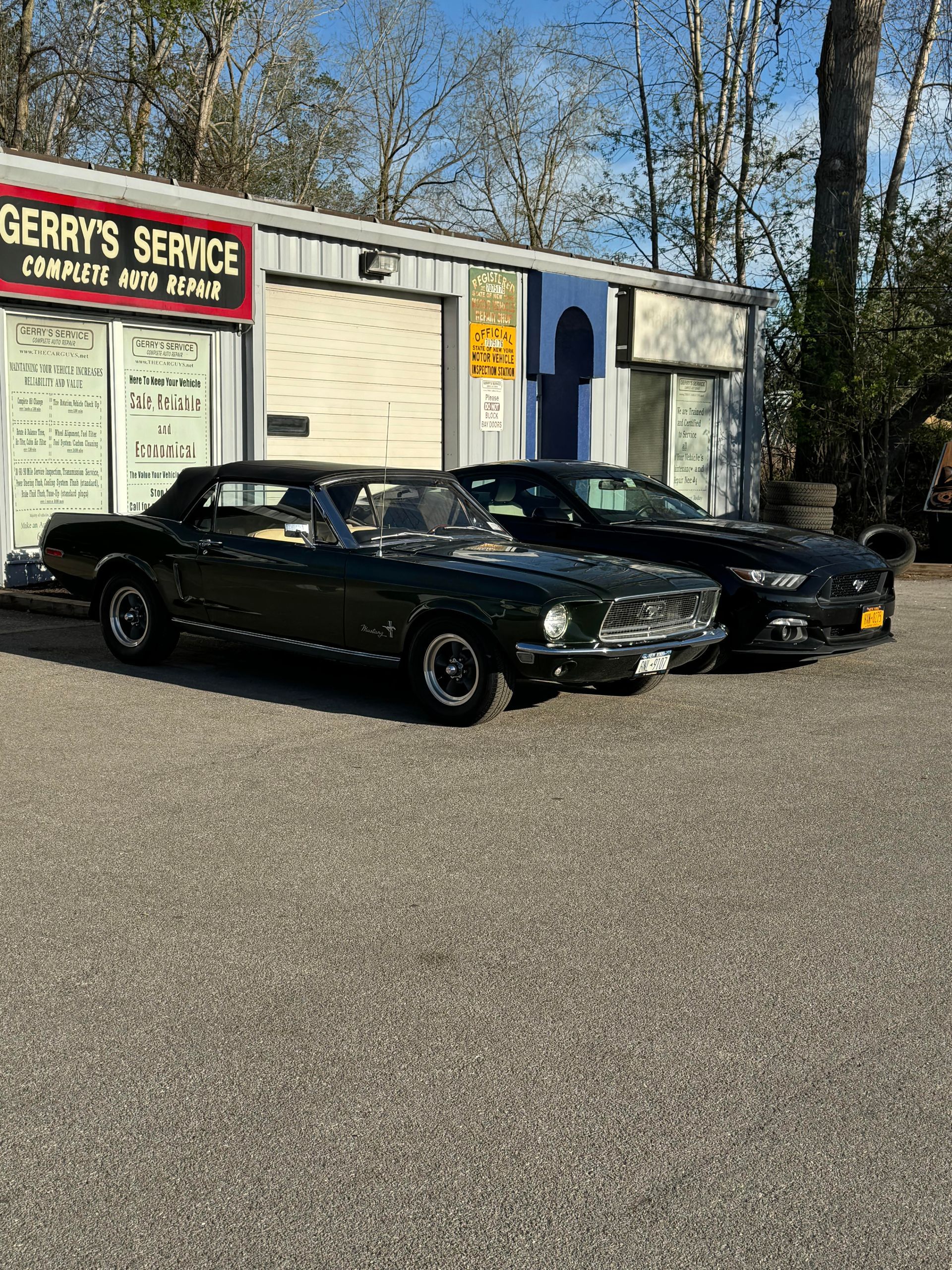 Two dark green Ford Mustangs parked in front of a service garage on a gravel lot.