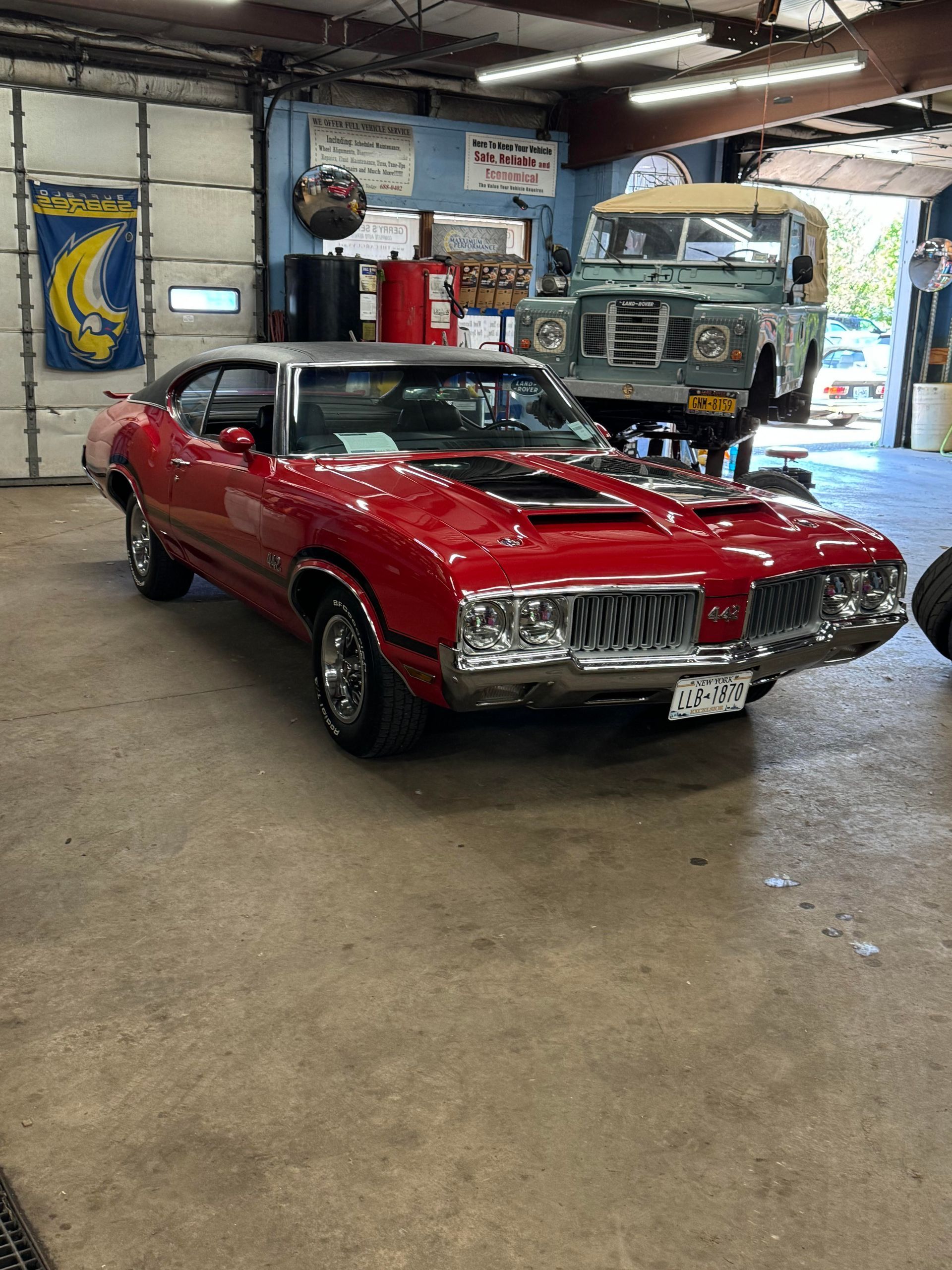 Red classic car in garage with green SUV in the background.