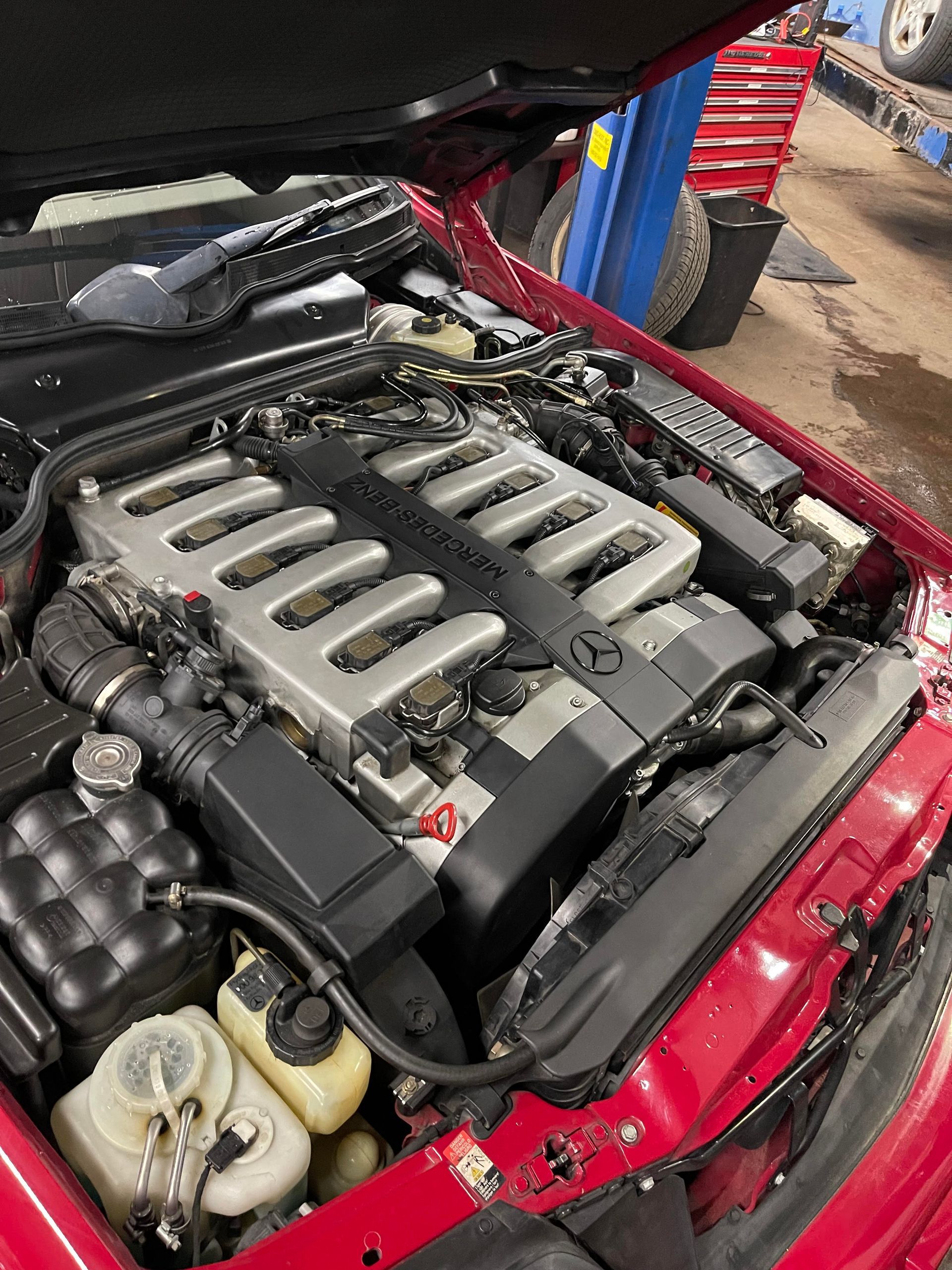 A red Mercedes engine bay with a large silver engine. The hood is up in a garage setting.