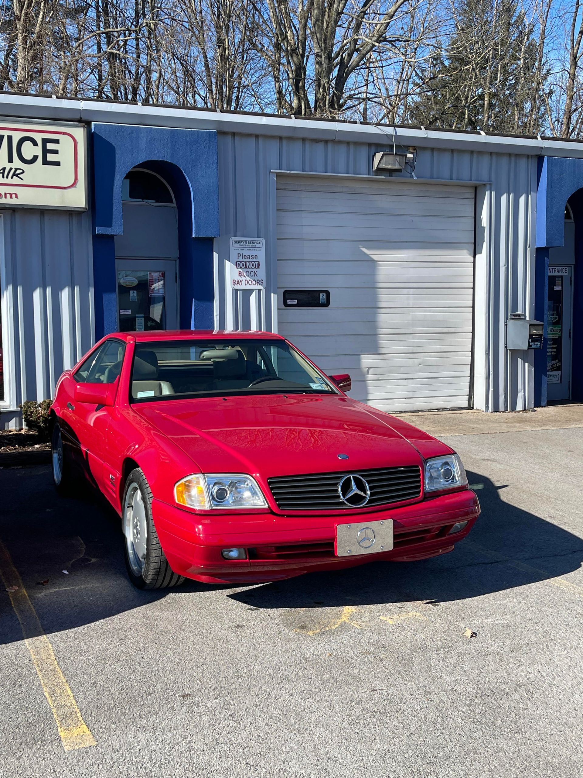Red Mercedes SL parked in front of a blue and gray auto shop with a closed garage door on a sunny day.