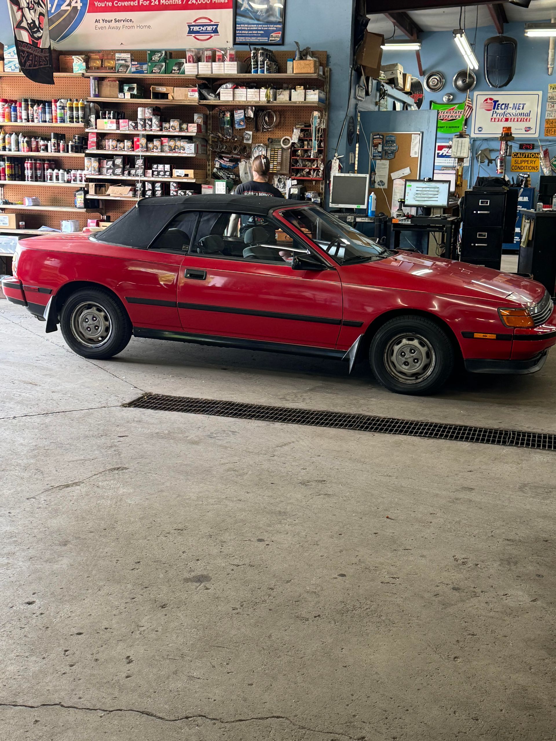 Red convertible car parked inside an auto shop. Black convertible top, silver wheels, and parts on shelves.