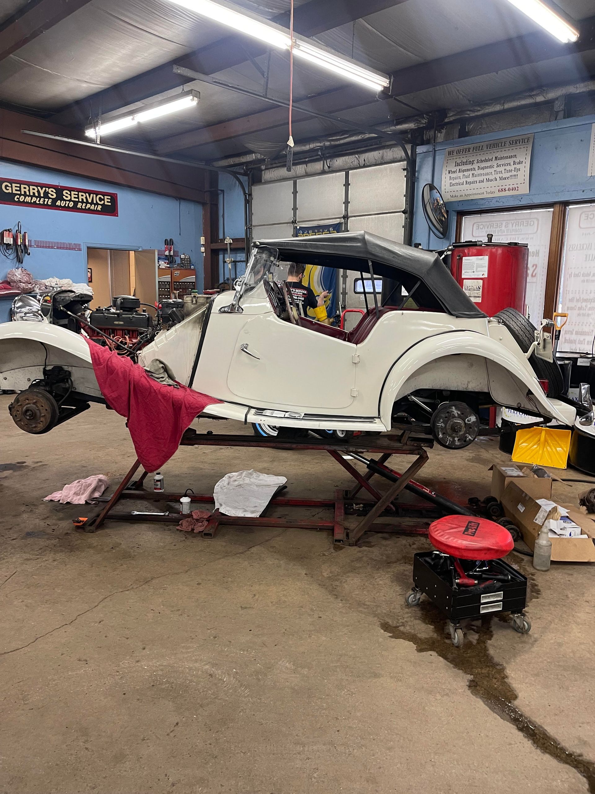 White car body lifted in a garage, undergoing repair. Red shop rag and tools visible.