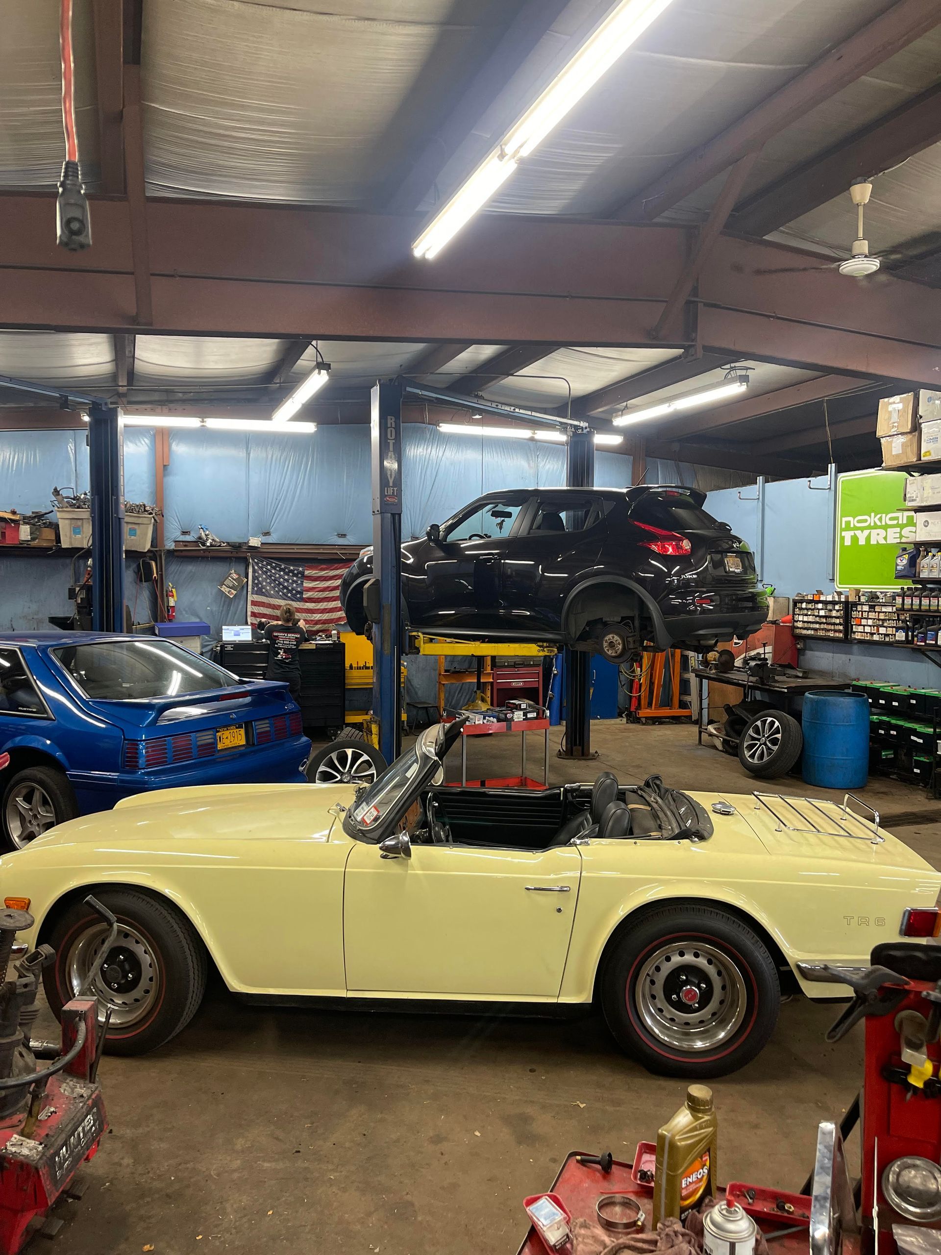 Yellow convertible in a car repair shop with a car on a lift and blue car in the background.
