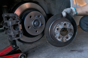 Person holding brake rotor next to a car's brake assembly; red jack in the background.