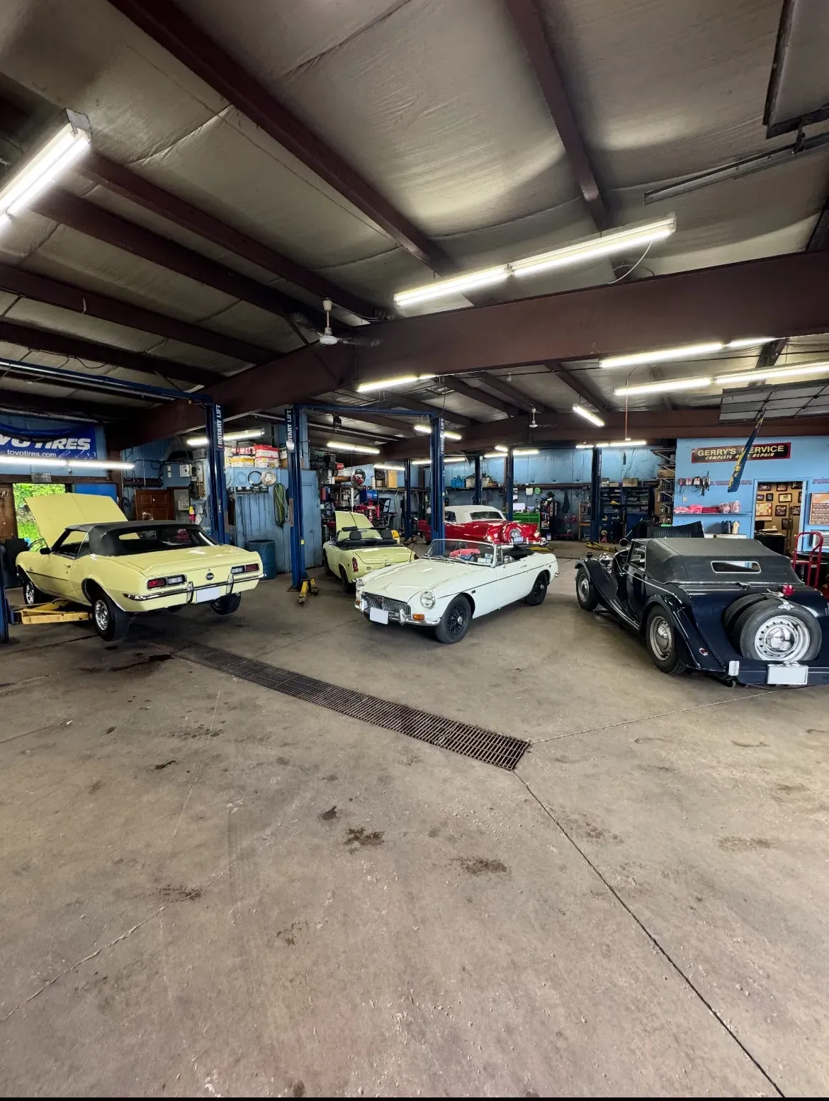 Classic cars in a garage. A yellow convertible has its hood up. Other cars are white, red, and dark blue.