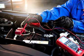 Mechanic in blue coveralls testing a car battery with a multimeter in a garage setting.
