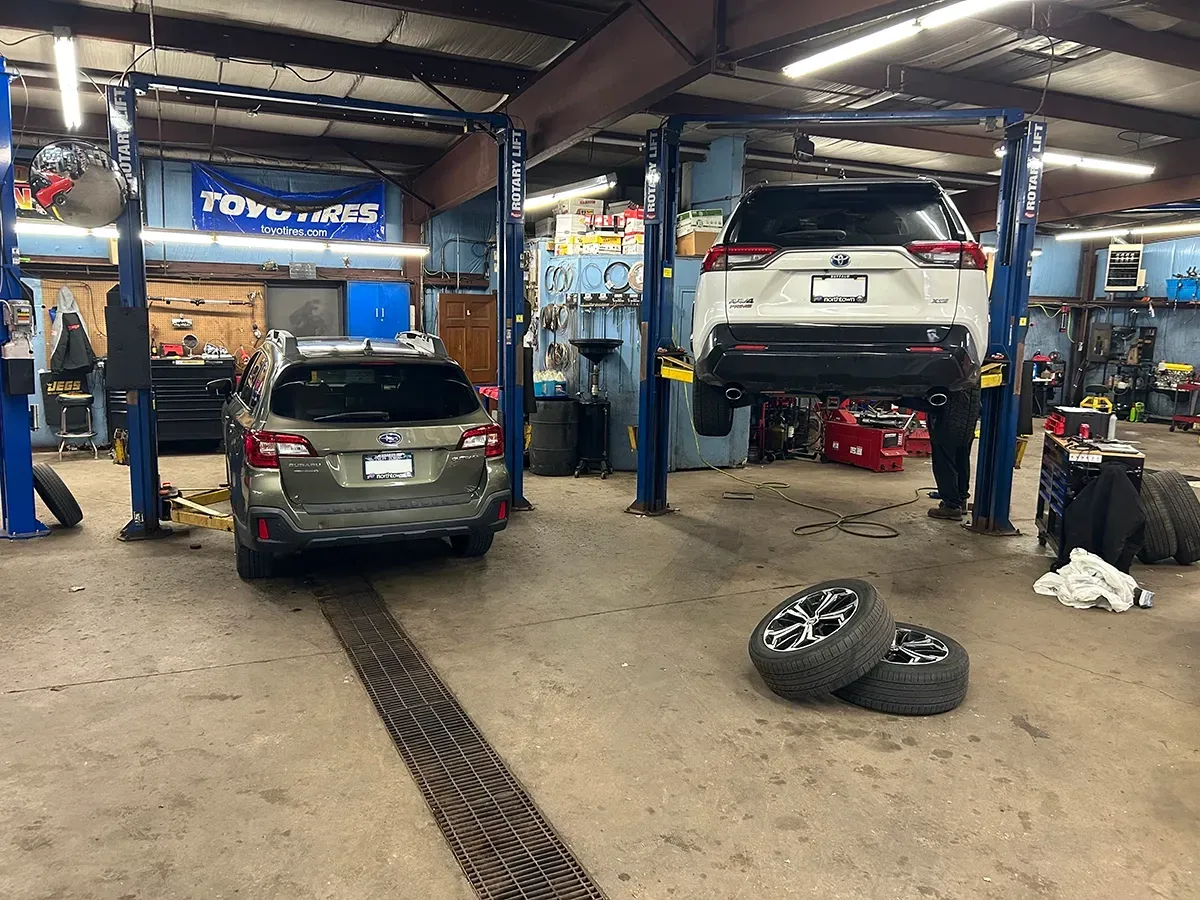 Two vehicles in a garage, one on a lift. Tools and tires scattered on the floor.
