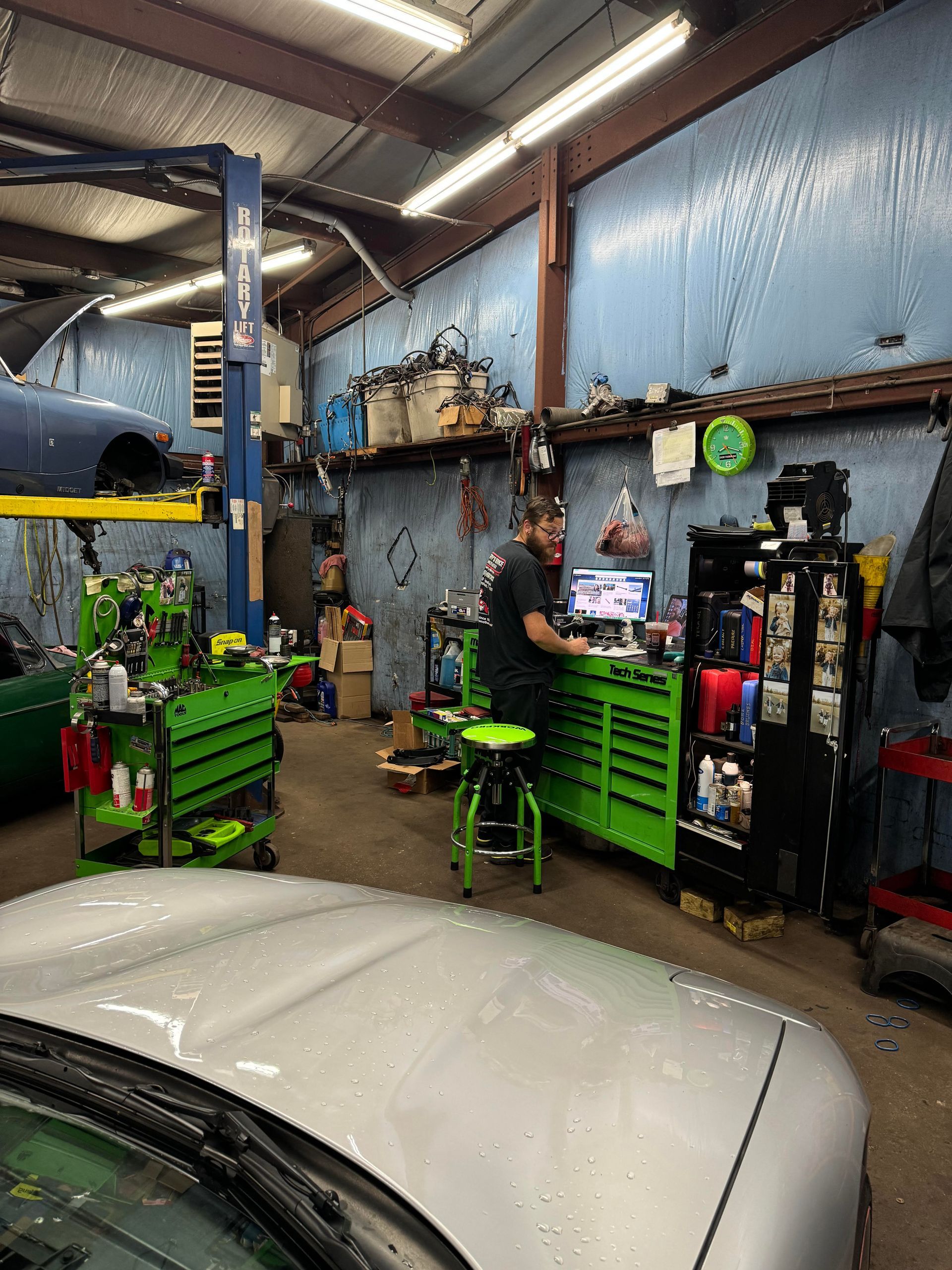 Mechanic working on a computer in a cluttered garage, silver car in foreground, green toolboxes, car on a lift.
