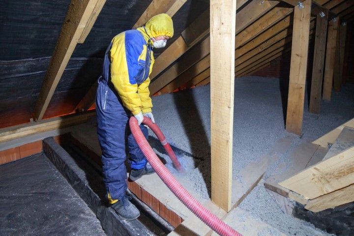 A worker in protective clothing uses a blower hose to install loose-fill insulation into an attic between wooden rafters.