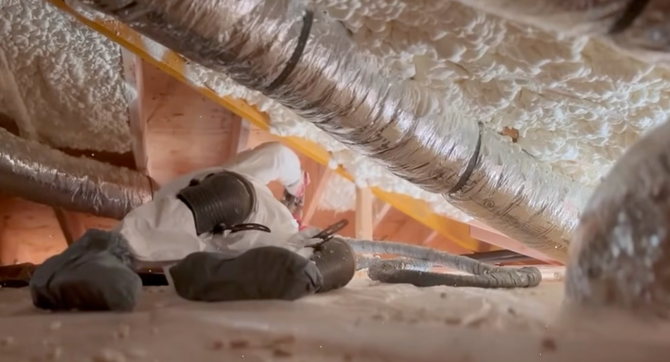 A person in protective gear working in a cramped attic space with foam insulation and flexible ductwork.