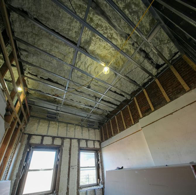A room under renovation with exposed ceiling metal framing, spray-foam insulation, wood wall studs, and two windows.