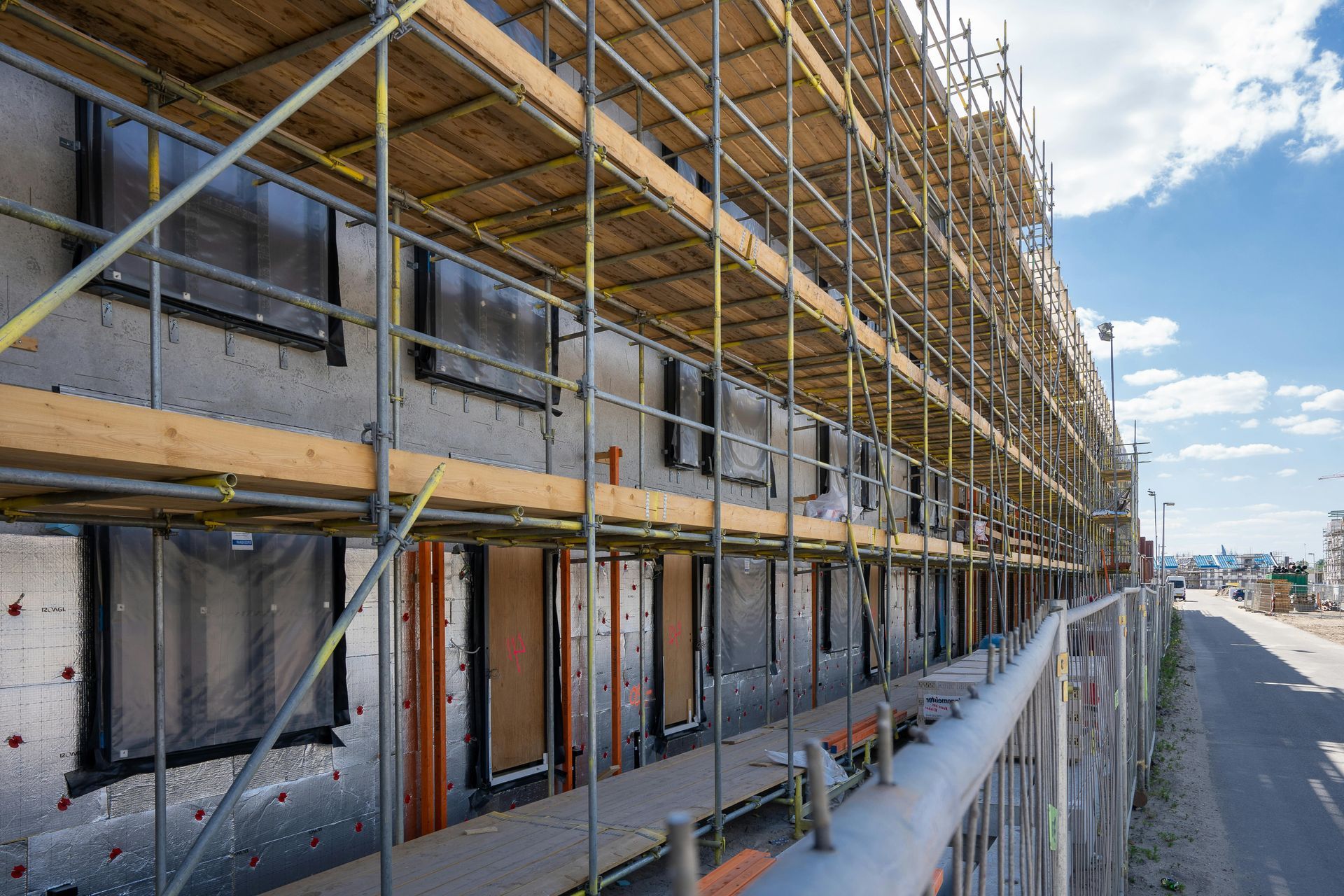 A building under construction with silver insulation panels, black window frames, and exterior scaffolding on a sunny day.