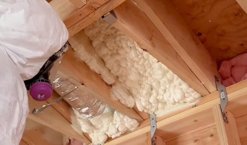 A person in a protective suit applies spray foam insulation between wooden ceiling joists.