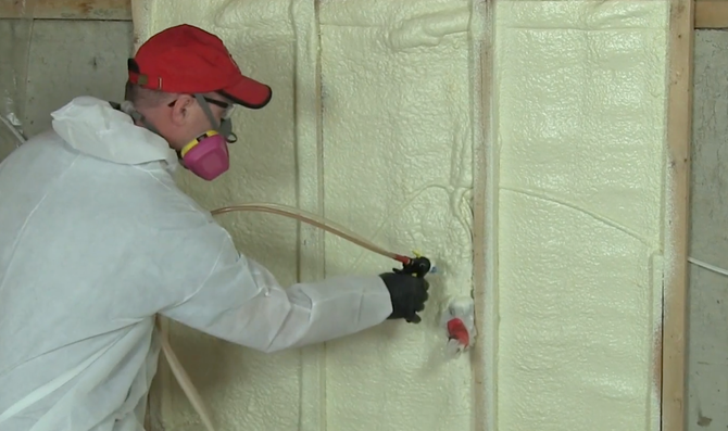 A person in protective gear and a respirator sprays foam insulation onto a wall between wooden studs.