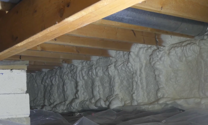 Crawl space foundation wall insulated with white spray foam, with wooden floor joists above and a vapor barrier below.